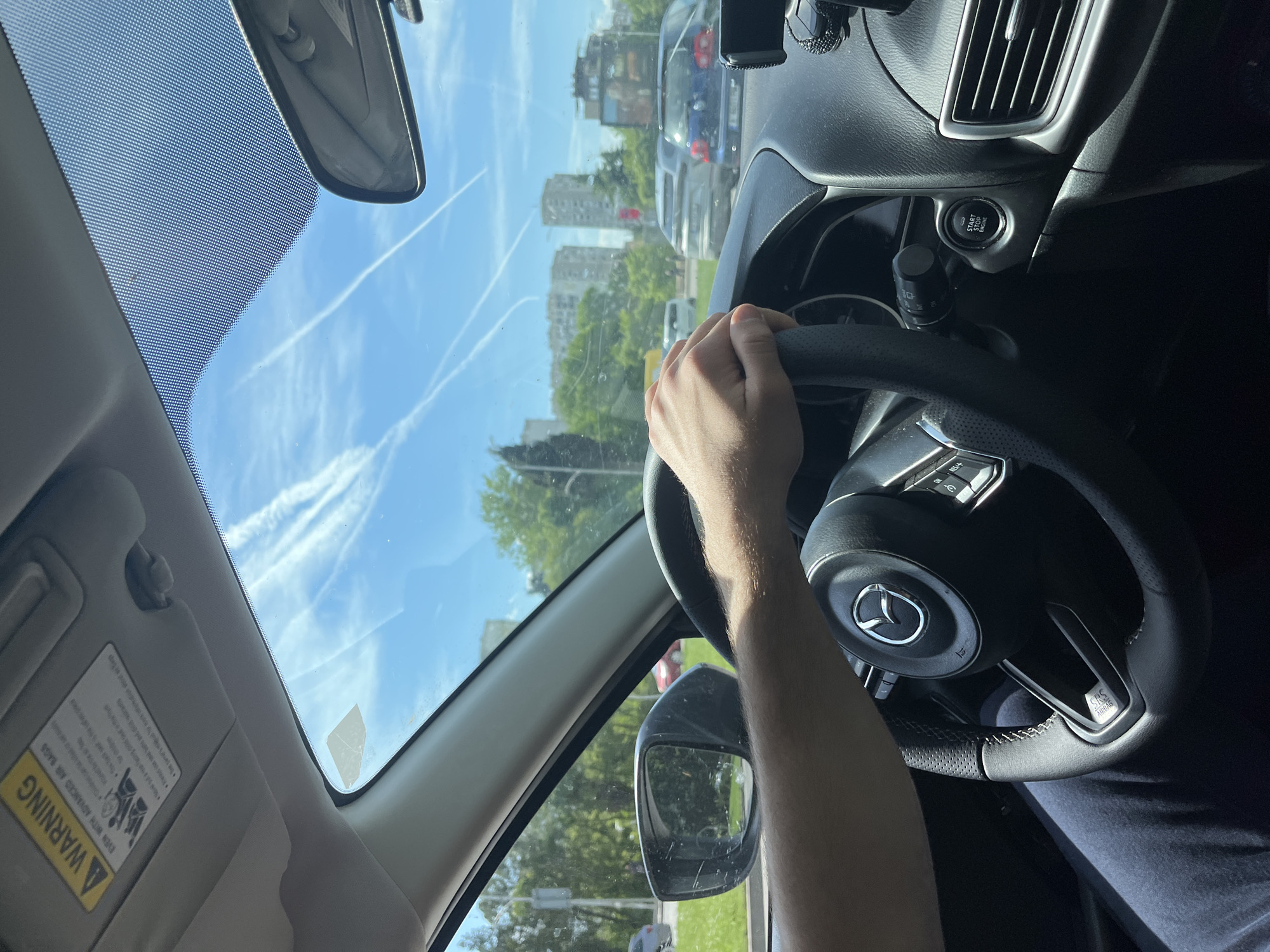 The photo shows the interior of a car, specifically the driver's side. The driver's left hand is visible on the steering wheel, which has a Mazda logo in the center. The dashboard, vents, and start/stop engine button are visible. Through the windshield, a blue sky with contrails can be seen, along with some buildings and trees, indicating a city environment. A warning label is on the sun visor. The rearview mirror is visible at the top of the windshield. Other cars are visible through the windshield. The location of the scene is an urban environment.
