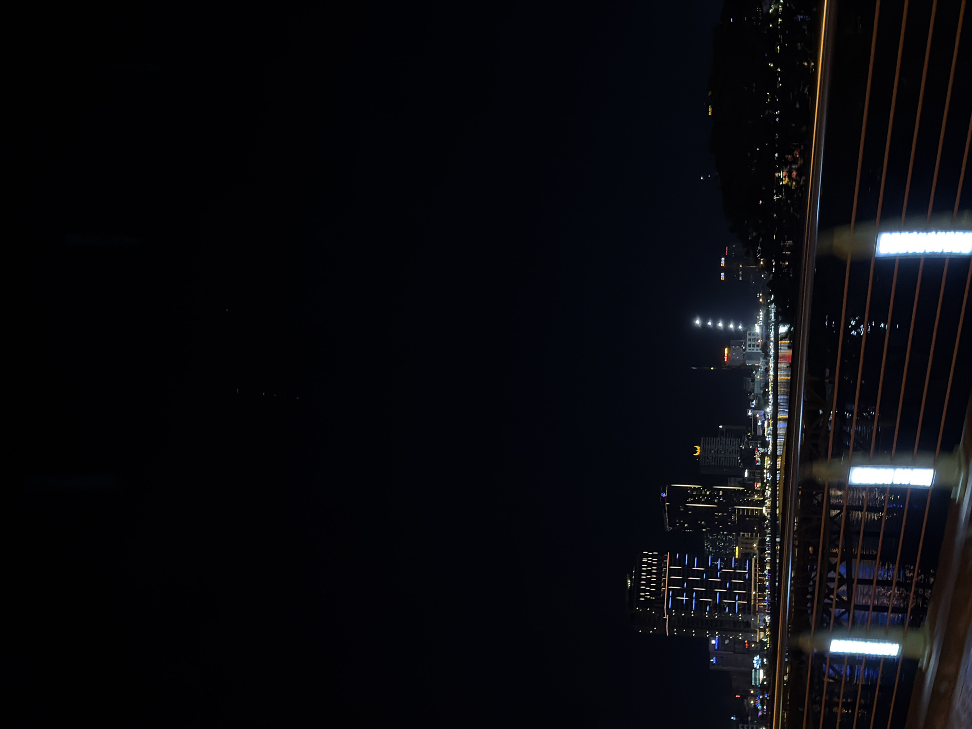 This is a night time photograph of a city skyline, likely taken from a bridge or elevated viewpoint in Da Nang, Vietnam. The image is oriented vertically.
The foreground features a railing with vertical wooden or metal bars, some of which are illuminated by bright white lights. Beyond the railing, the dark expanse of a body of water reflects the lights of the city.
The main subject is the brightly lit cityscape in the distance. Several modern buildings of varying heights are visible, adorned with numerous illuminated windows. Some buildings feature distinctive patterns of lights, such as blue grid-like designs on one prominent structure. The lights create a vibrant and lively atmosphere, contrasting with the dark night sky. The skyline stretches across the horizon, hinting at a bustling urban environment.
The time of day is clearly night, evidenced by the darkness and the artificial illumination of the city. The weather appears clear, allowing for a crisp view of the city lights and their reflections. The presence of tall, modern buildings suggests an urban setting, and the visual cues, combined with the provided location context, point to Da Nang.
There is no discernible text visible in the image. The overall mood conveyed is one of urban dynamism and the beauty of a city at night.