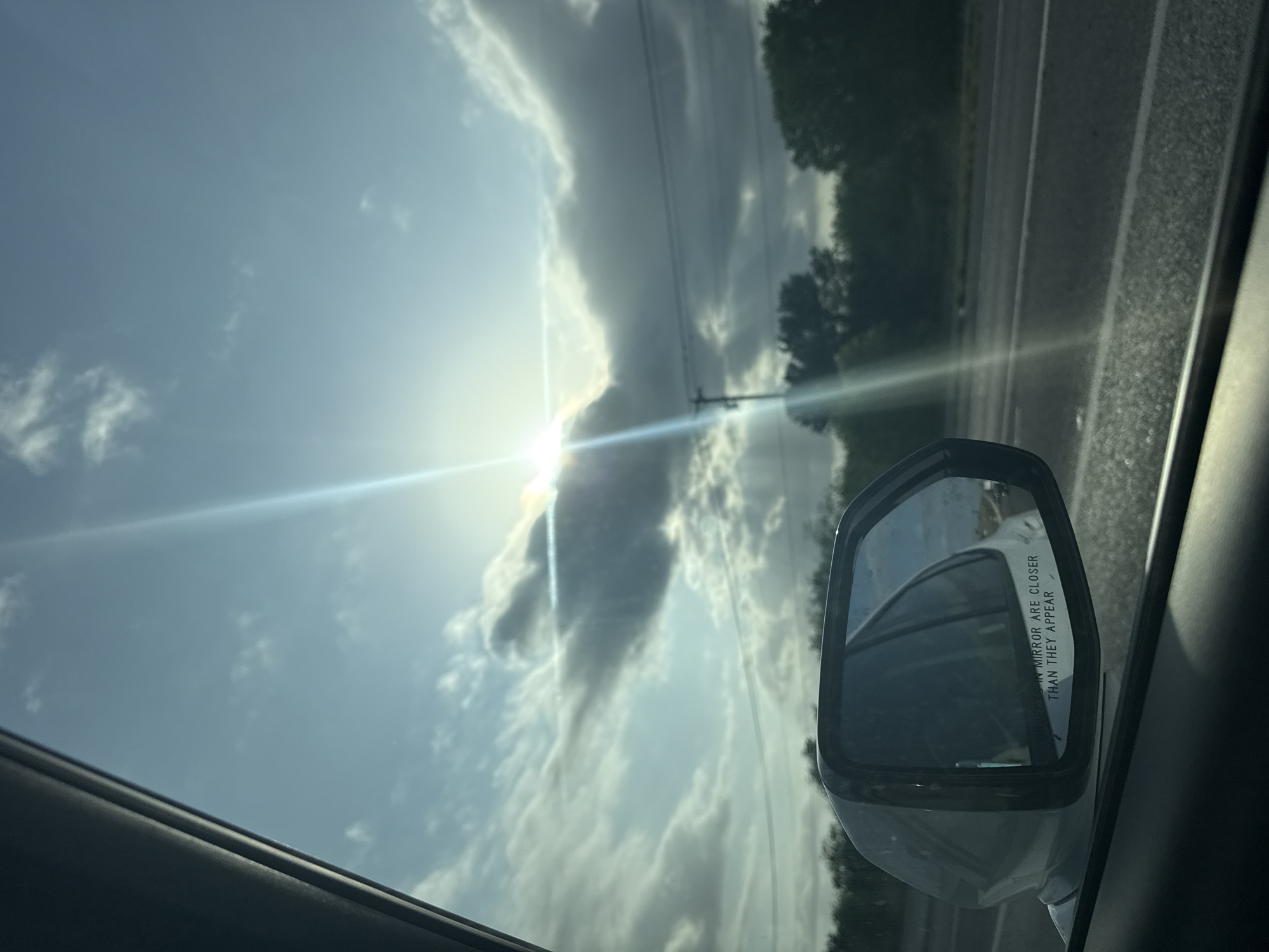 The image shows a view from inside a car. The sky is visible, with scattered clouds and the sun shining brightly. There is a side-view mirror in the foreground, reflecting the scenery behind the car, including the road and some trees. The scene appears to be taken while driving on a road or highway, evidenced by the movement suggested and the road visible outside the car. The location in New Braunfels, United States of America, cannot be determined.