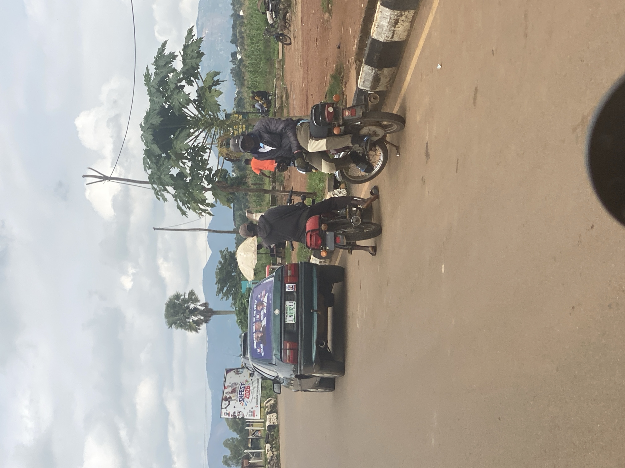 The image depicts a street scene in Maigizo, Nigeria, under an overcast sky. In the foreground, a green sedan with a license plate "NJN-897AJ" is parked on the left side of the road. The car's rear window is adorned with several stickers, one of which reads "NIGERIA WILL BE OK 2023". Two individuals are seated on motorcycles on the right side of the road, facing away from the camera. The motorcycle closest to the viewer has a red rear fender. Further down the road, a billboard with "SKFEST 2023" and images of people is visible on the left. Trees, including a palm tree and a papaya tree, are present in the background, along with hills and farmland. Other parked motorcycles and some people can be seen in the distance. The road is paved, with a curb marked by black and white stripes.