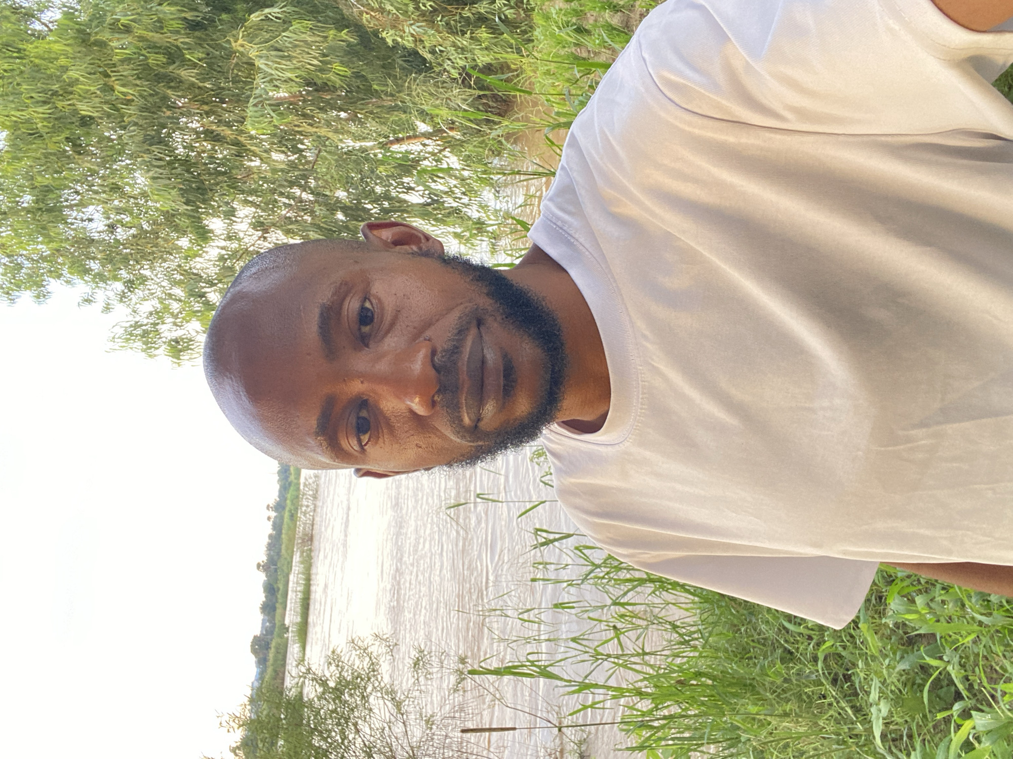 A close-up, eye-level, medium shot captures a Black man with a shaved head and a short beard. He is positioned in the foreground, facing forward and looking directly at the camera. He is wearing a white t-shirt. The background features a body of water, likely a river, with lush green vegetation, including tall grasses and trees, bordering its edge. In the distance, across the water, a line of trees and possibly some structures suggest a settlement. The overall lighting indicates it is daytime, possibly late afternoon or early morning, given the soft, diffused light. The scene appears to be outdoors in a natural environment. The provided crop hints suggest the location is near a river in Rido, Nigeria.