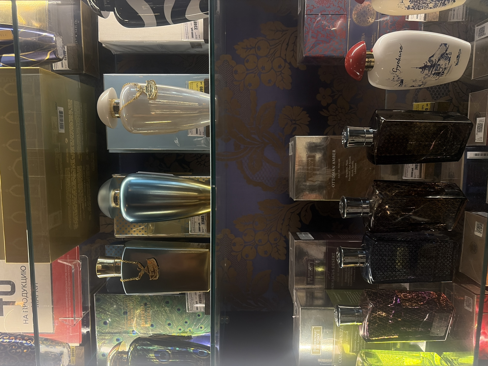This media file is a photograph of a retail display case, likely in a department store or duty-free shop, showcasing a variety of perfumes. The display is well-lit and organized, with perfumes arranged on multiple shelves.
Prominent objects include several perfume bottles of various shapes, sizes, and colors. Some are clear glass, allowing the colorful liquid inside to be seen, while others are opaque or have patterned designs. Notable among these are a tall, iridescent blue bottle and a cream-colored bottle with a gold chain and a red cap. Several rectangular perfume boxes with intricate designs are also visible, some featuring floral patterns and others with what appears to be a snake-skin texture.
The background of the display case has a decorative pattern, possibly wallpaper or fabric, with large, stylized yellow floral motifs against a dark purple background. There are price tags and labels visible on some of the items, with some text in Russian, indicating the location might be in a Russian-speaking region. One tag reads "НА ПРОДУКЦИЮ" and another "29980 руб." indicating a price and currency. Another label visible on a box says "THE MERCHANT OF VENICE".
The scene appears to be an indoor retail environment, with no indication of time of day or weather. The overall impression is one of a curated and appealing product display, designed to attract customers. The focus is entirely on the products themselves, with no people present.