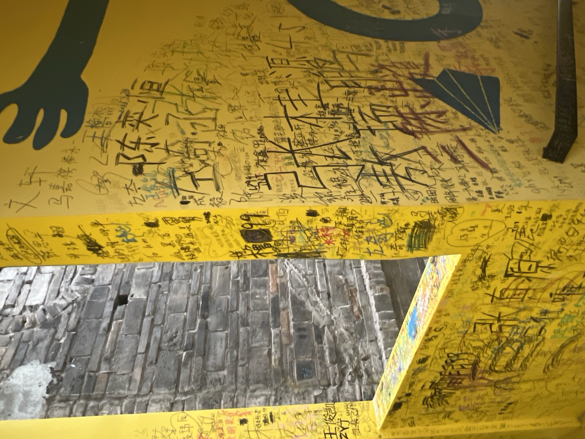 The image shows a yellow wall covered in graffiti. The graffiti includes Chinese characters, numbers, and drawings. In the background, a stone wall is visible. The location is identified as Yuzhong District, China. The graffiti appears to be a form of public art or expression.