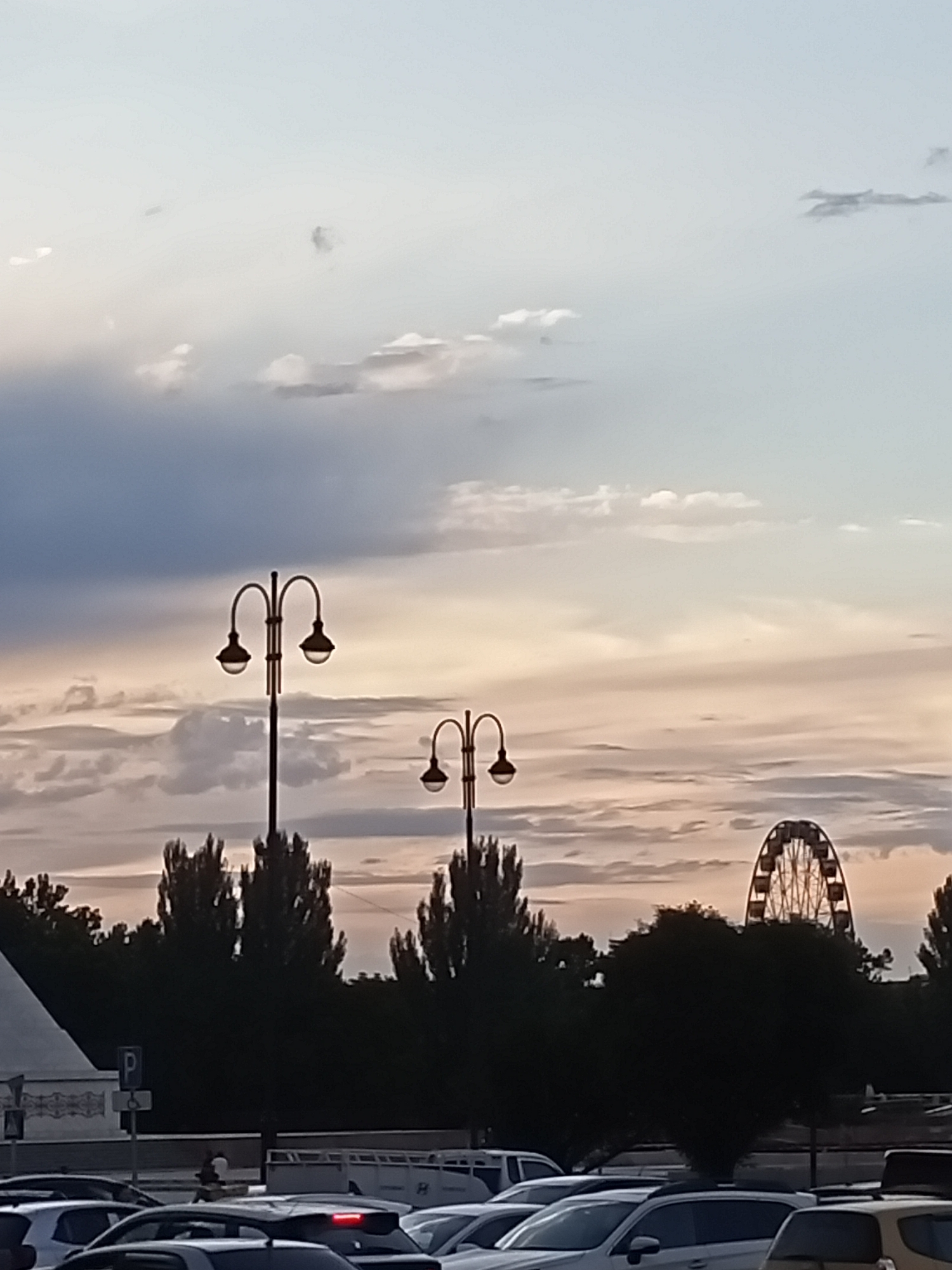 The image depicts a street scene at dusk, with a sky filled with clouds in shades of gray, white, and a hint of orange and pink. Two decorative streetlights with arched arms stand prominently in the foreground, silhouetted against the sky. In the middle ground, a row of dark, tall trees partially obscures a Ferris wheel in the distance, which is illuminated by lights. The lower portion of the image shows a street with several cars parked and driving, indicating an urban setting. There are also hints of buildings and infrastructure, including a white structure on the far left and a parking sign. The overall impression is a somewhat hazy, possibly humid, evening in a city environment. The location is identified as Bishkek City, Kyrgyzstan.