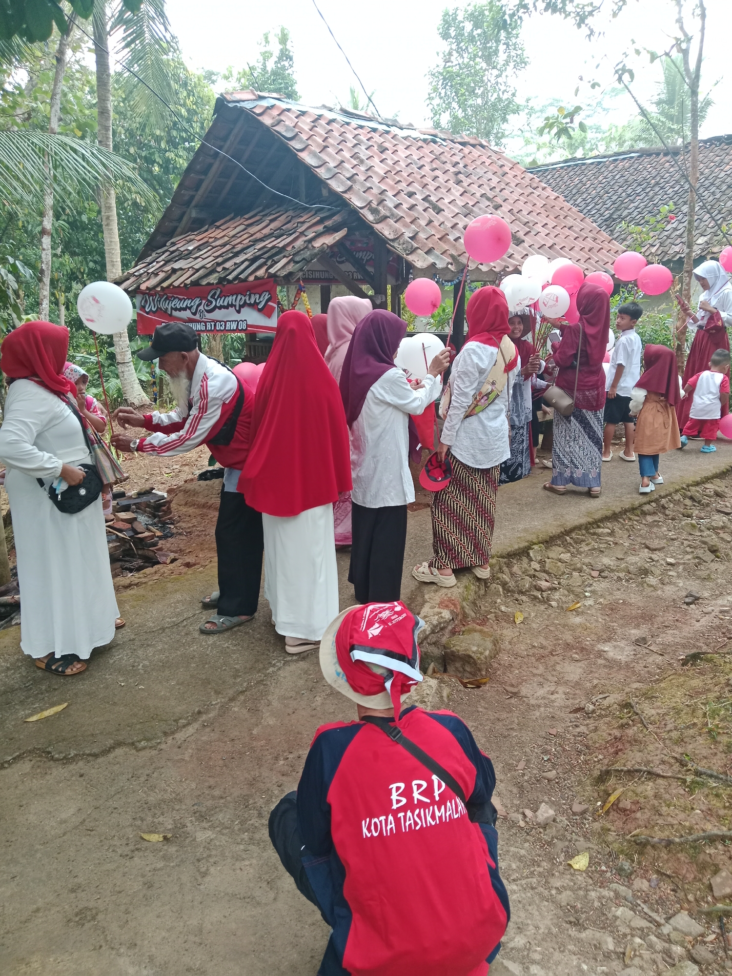 A group of people, predominantly women and children, are gathered outdoors in Bojongmengger, Indonesia, near a building with a tiled roof. Many of the women are wearing headscarves and long dresses. Some are holding balloons, which are pink and white. A man with a white and red jacket and a black cap is interacting with a woman in a white dress. In the foreground, a person in a red shirt with "BRP KOTA TASIKMALALAYA" written on it is partially visible, crouched down. The ground is a dirt path with some rocks and vegetation. The overall atmosphere appears to be a community gathering or event.