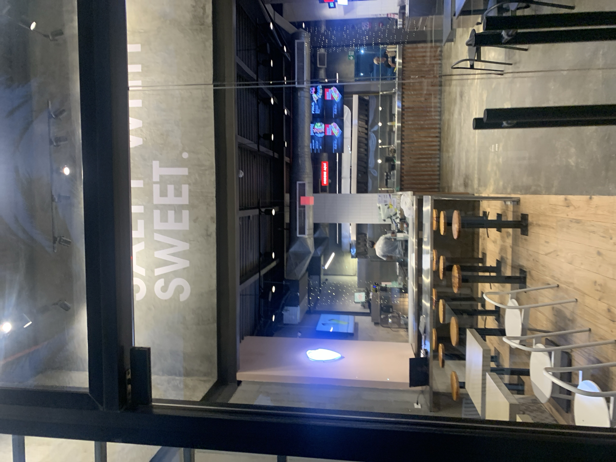 The image presents an interior view of a modern commercial establishment, likely a cafe or restaurant, seen through a large glass window or partition. The scene is illuminated by artificial lighting, suggesting it is nighttime or evening.
The interior design features an industrial aesthetic with exposed dark ceiling structures and various spotlights. On a concrete-colored wall to the left, large white letters are visible, spelling "SALT" and clearly "SWEET".
Inside, the space is brightly lit. There are multiple seating arrangements, including a counter with round wooden stools on black bases, and several light-colored rectangular tables accompanied by white chairs. Towards the back, a service area is equipped with digital menu screens displaying food items and the Spanish text "ORDENA AQUI" (Order Here). At least two individuals are present within the establishment: one person is seen behind a counter, appearing to work, and another is further in the background. A prominent glowing blue, oval-shaped sign or logo is positioned on a low, light-colored platform in the middle ground. The flooring transitions from wooden planks in the seating area to what appears to be a concrete or tile surface elsewhere. Reflections are visible on the glass surface, particularly on the left side.
