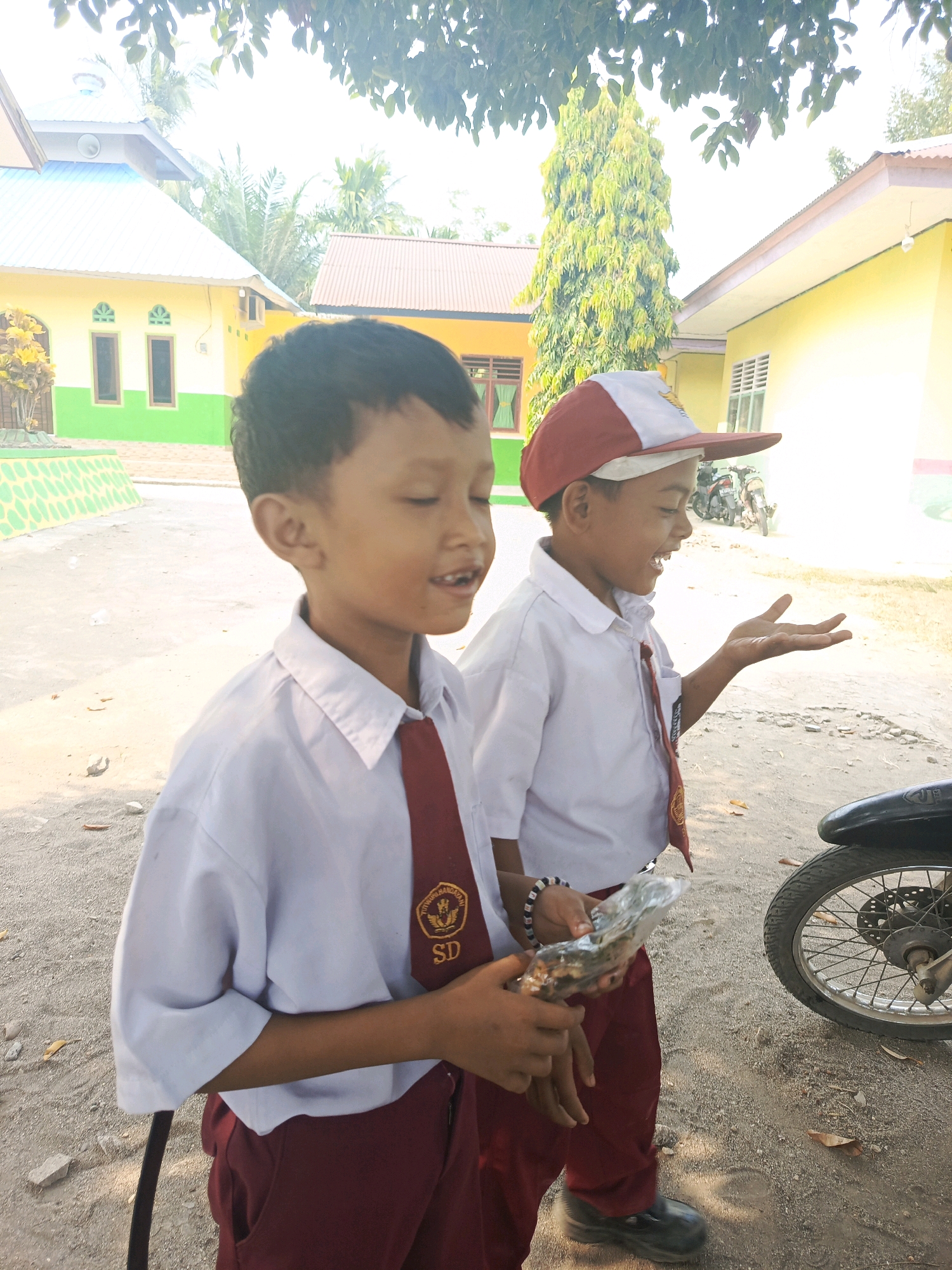 Two young boys, dressed in school uniforms consisting of white shirts, red ties, and maroon pants, stand outside. The boy on the left holds a clear plastic bag containing food. The boy on the right wears a red and white cap and gestures with his open hand. In the background, two yellow buildings with green and white accents are visible, along with trees and a motorcycle parked near the right edge of the frame. The ground is covered in gravel and scattered leaves. This scene is located in Karya Dadi, Indonesia.