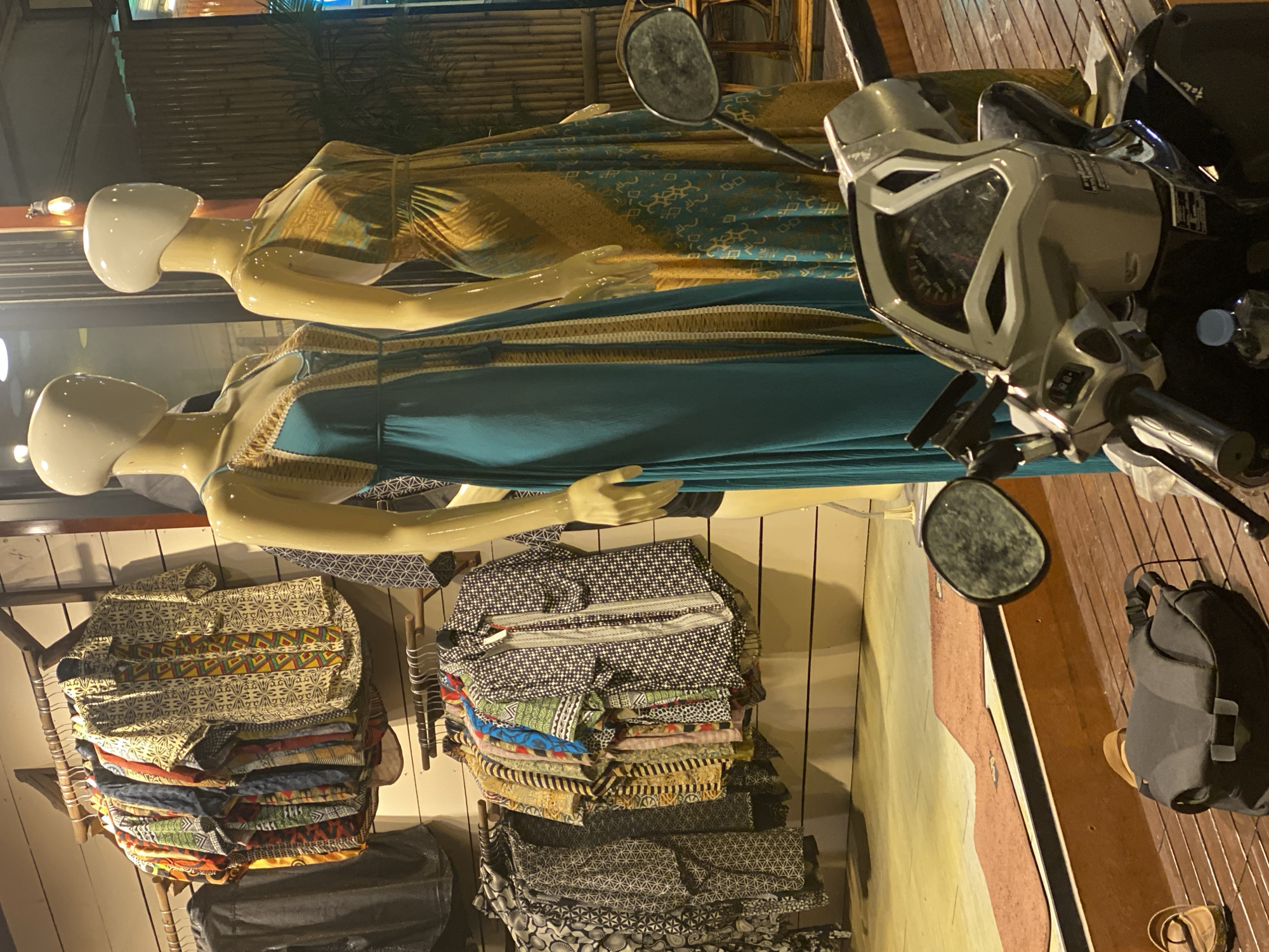 Two mannequins displaying dresses stand in front of a storefront in Baan Kai, Thailand. The mannequins are faceless with smooth, white heads and yellow-tinted bodies. The top mannequin is wearing a yellow dress with a blue pattern, while the bottom one is wearing a blue dress with golden accents. In front of the mannequins is a motorcycle, with its mirror partially obstructing the view of the top mannequin. Behind the mannequins, there are several racks of folded shirts with various patterns, hanging against a light-colored wall. The store appears to be located outdoors, possibly on a wooden deck. A backpack and a pair of sandals are visible next to the motorcycle.