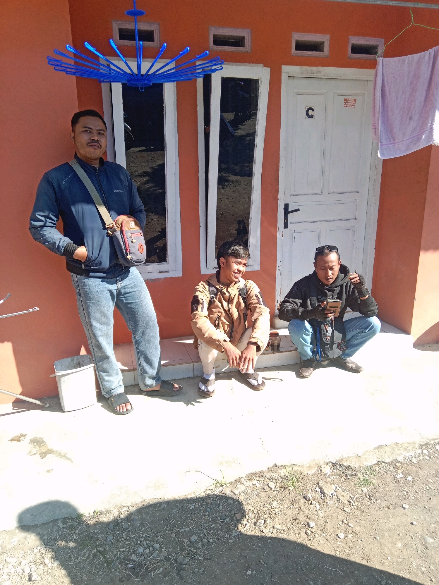 The image depicts three adult males standing and squatting in front of a building with an orange facade and white doors and window frames. A blue plastic drying rack is suspended above and to the left of the entrance. A white towel is hanging on the right side of the building.
The male on the far left is standing and wearing a dark blue jacket, light blue jeans, and black sandals. He has a cross-body bag over his right shoulder. His left hand is in his jeans pocket. He has a mustache and short dark hair.
The male in the center is squatting and wearing a tan jacket with dark detailing, beige pants, and flip-flops. He is smiling and looking to his left.
The male on the far right is squatting and wearing a dark gray hooded sweatshirt, blue jeans, and dark shoes. He has sunglasses on his head and is wearing black fingerless gloves. He is holding a mobile phone in his hands and appears to be looking at it.
A white bucket is on the ground to the left of the standing man. Two cups are on the ground to the left of the squatting men. The ground in front of the building is paved. The scene appears to be outdoors, likely in a residential or commercial area. The presence of a building with numbered doors and outdoor clothing drying suggests a residential context. The overall lighting and shadows indicate it is daytime.
The location is described as being in the city of Leksono, Indonesia.