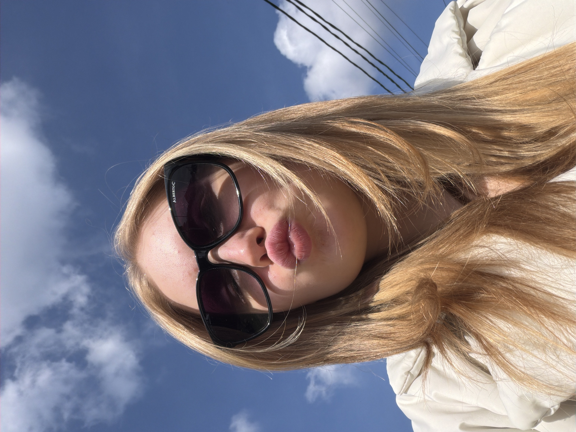An outdoor, low-angle portrait captures a young individual with long, layered blonde hair and a light-colored puffy jacket, set against a bright blue sky with scattered white clouds. The individual wears black-framed sunglasses, bearing the inscription "ALBERTO C" on the right temple arm, and has their lips pursed into a "kiss" expression. Strong natural light casts shadows from the sunglasses. Three parallel utility lines are visible in the lower right background. The scene is located in the city of Rossiyskiy, Russia.