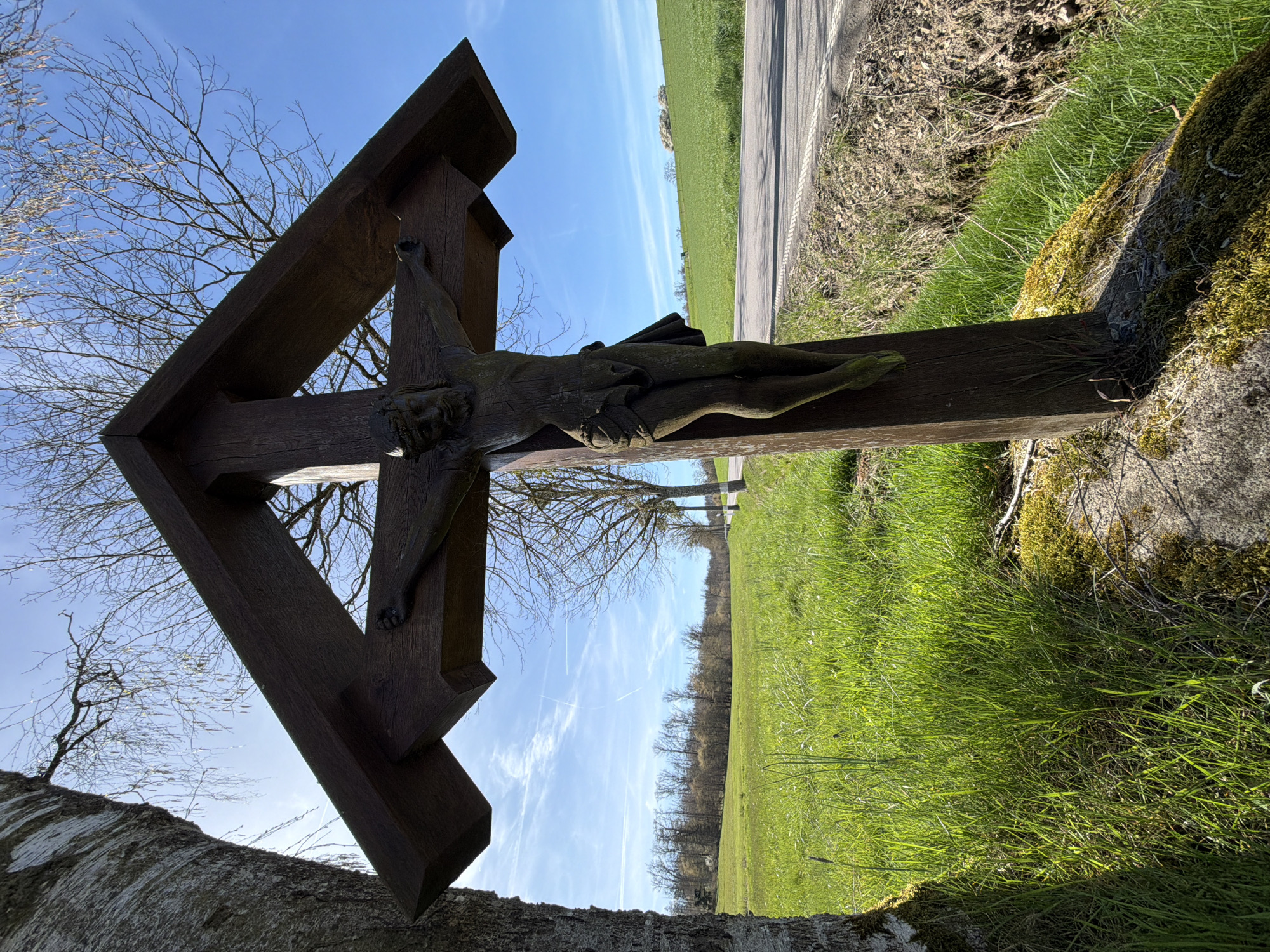 A wooden crucifix stands near a green field and a road, under a partly cloudy blue sky. Bare branches of trees are visible in the background. The cross is placed on a moss-covered stone or structure. Based on visual cues, it is likely located near Junglinster, Luxembourg.