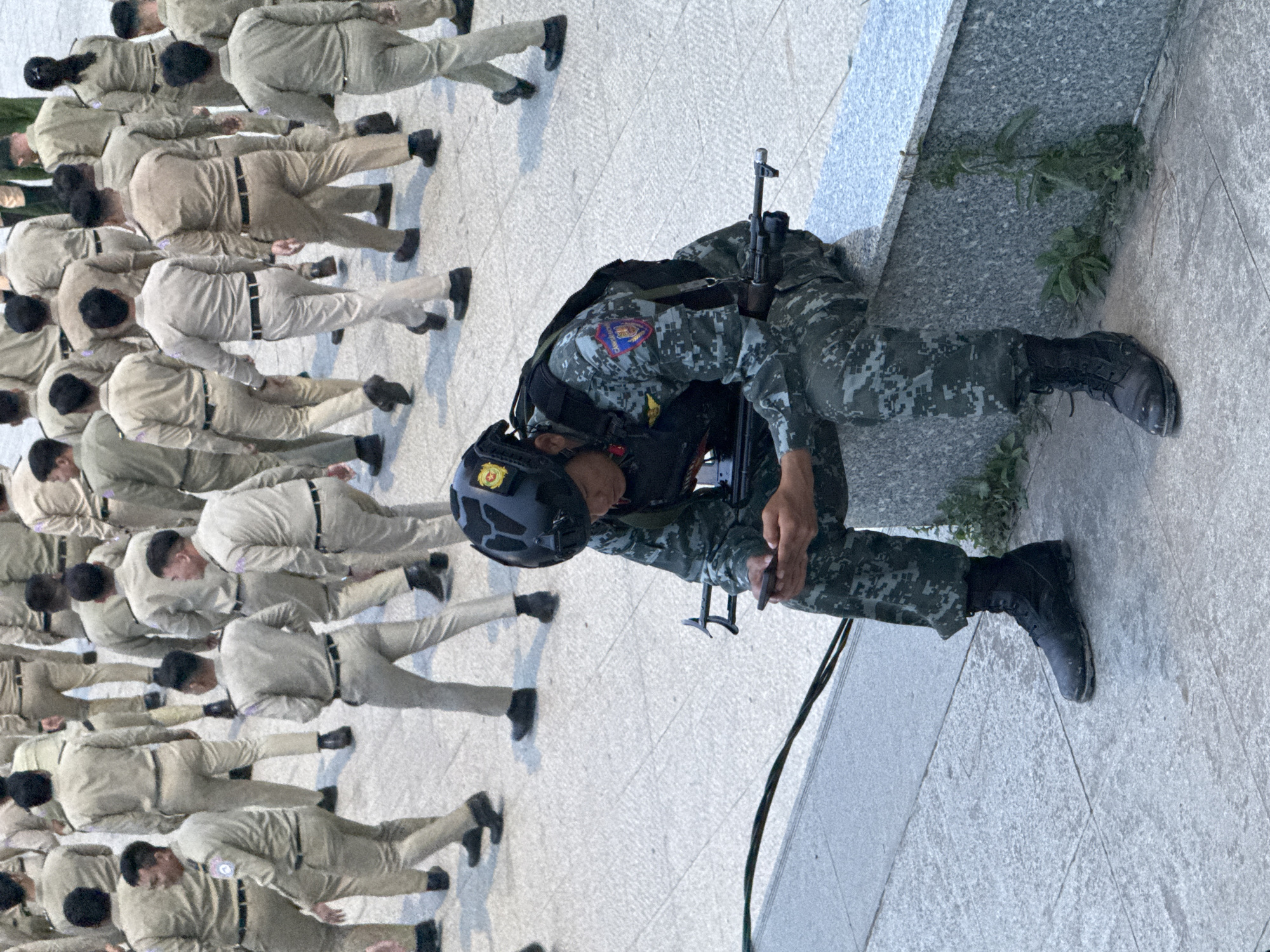 This image depicts a group of people, likely cadets or trainees, in uniform standing or kneeling in a line on a paved outdoor area. The majority of the individuals are wearing light-colored uniforms with black belts and black shoes. One person, distinctively dressed in camouflage tactical gear, a helmet, and holding a rifle, is kneeling to the right of the frame. This individual appears to be in a position of attention or perhaps undergoing some form of training or drill.
The setting appears to be an open courtyard or training ground, characterized by the light-colored paving stones and a low, grey granite wall with some greenery growing along its base. The lighting suggests it could be daytime, with the sun casting shadows.
The overall mood is one of discipline and order, with the uniformed individuals standing in formation. The presence of the armed individual with tactical gear suggests a potential military or law enforcement training context. There are no discernible text elements in the image. The location context is Da Lat, Vietnam.