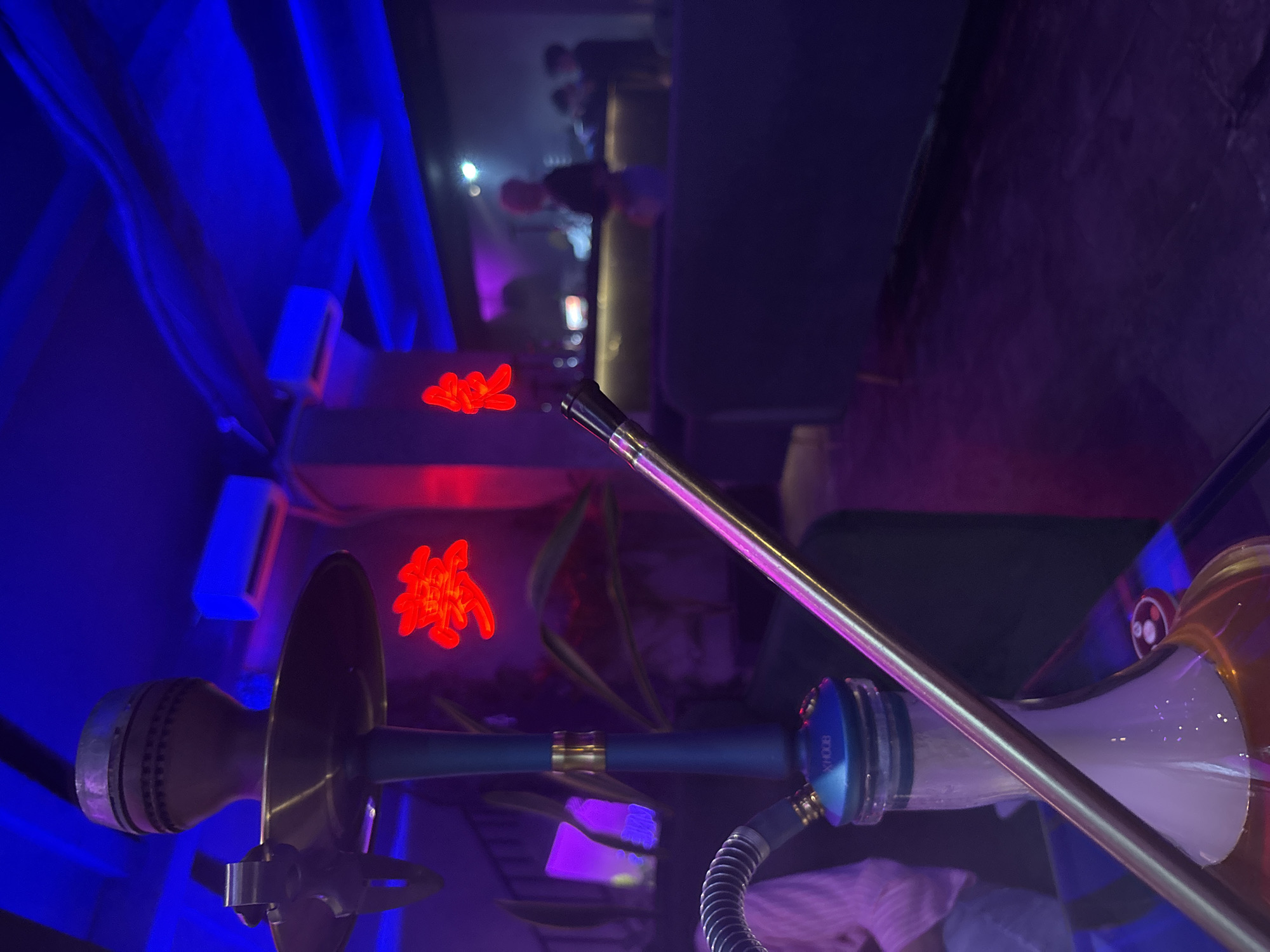 This image captures an indoor scene, likely a bar or shisha lounge, at night or in the evening. The foreground is dominated by a modern hookah, featuring a metallic stem, a glass base partially filled with liquid, and a hose connection.
The setting is characterized by vibrant, artificial lighting, with strong blue illumination on the left and foreground, contrasting with red and purple hues in the background. This creates a moody and atmospheric environment. In the blurry background, several indistinct figures of people can be seen, suggesting a social gathering or patrons in the lounge.
A prominent detail in the mid-ground is a glowing red neon sign displaying Chinese characters, which appear to be "囍" (Shuāngxǐ), symbolizing "double happiness." Other glowing blue light fixtures are visible along the walls. The overall activity seems to be leisurely socialization, with the hookah indicating shisha consumption. The location context of Kuta Selatan, Indonesia, is consistent with such a nightlife establishment. Faintly visible text "XHOOB" is present on a blue component of the hookah. The image is tilted, presenting a dynamic, angled view of the scene.