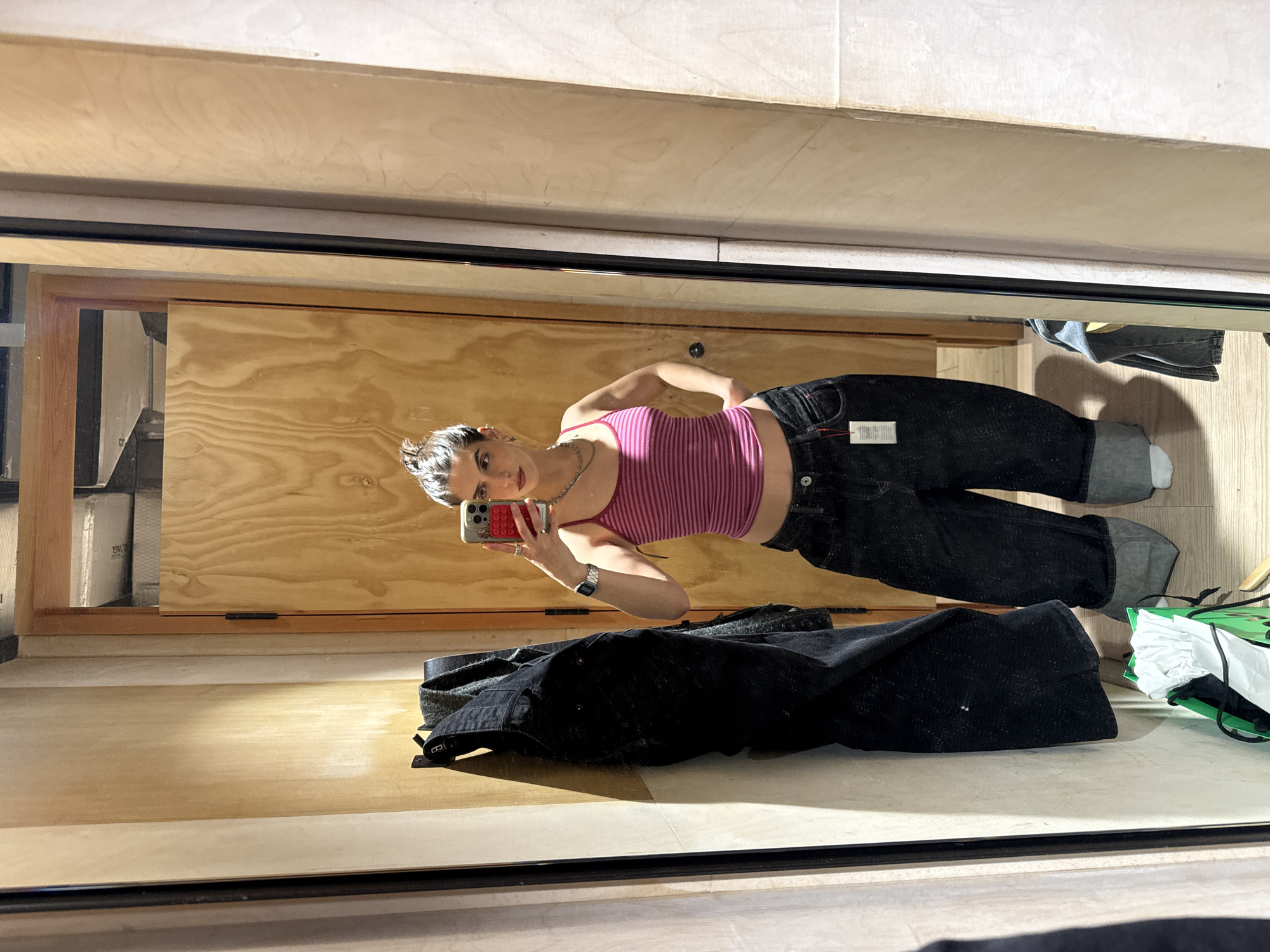 A young woman takes a mirror selfie inside a retail dressing room. The woman is wearing a pink and red striped tank top and dark wash jeans. She is holding a red phone in her left hand. A pair of jeans is on the floor. A shopping bag can be seen on the right side of the mirror. Based on the brand's Instagram posts and the style of the dressing room, the store is in Madrid, Spain.