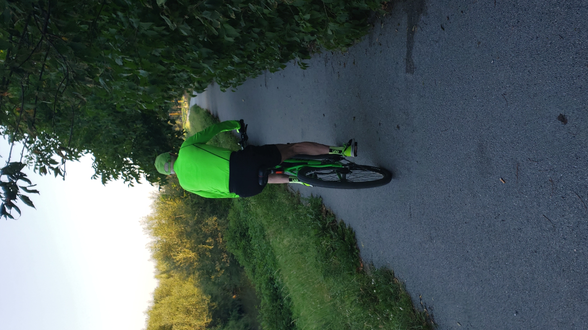 A man is riding a bicycle on a paved path bordered by greenery. He is wearing a bright green long-sleeved shirt, black shorts, a green cap, green socks, and black and green athletic shoes. The bicycle is black and green. The path appears to be a recreational trail, possibly along a river given the dense foliage and vegetation to the left of the path. The setting suggests an outdoor environment, likely during daylight. The image does not provide specific visual cues to pinpoint the location within Prešov, Slovakia.