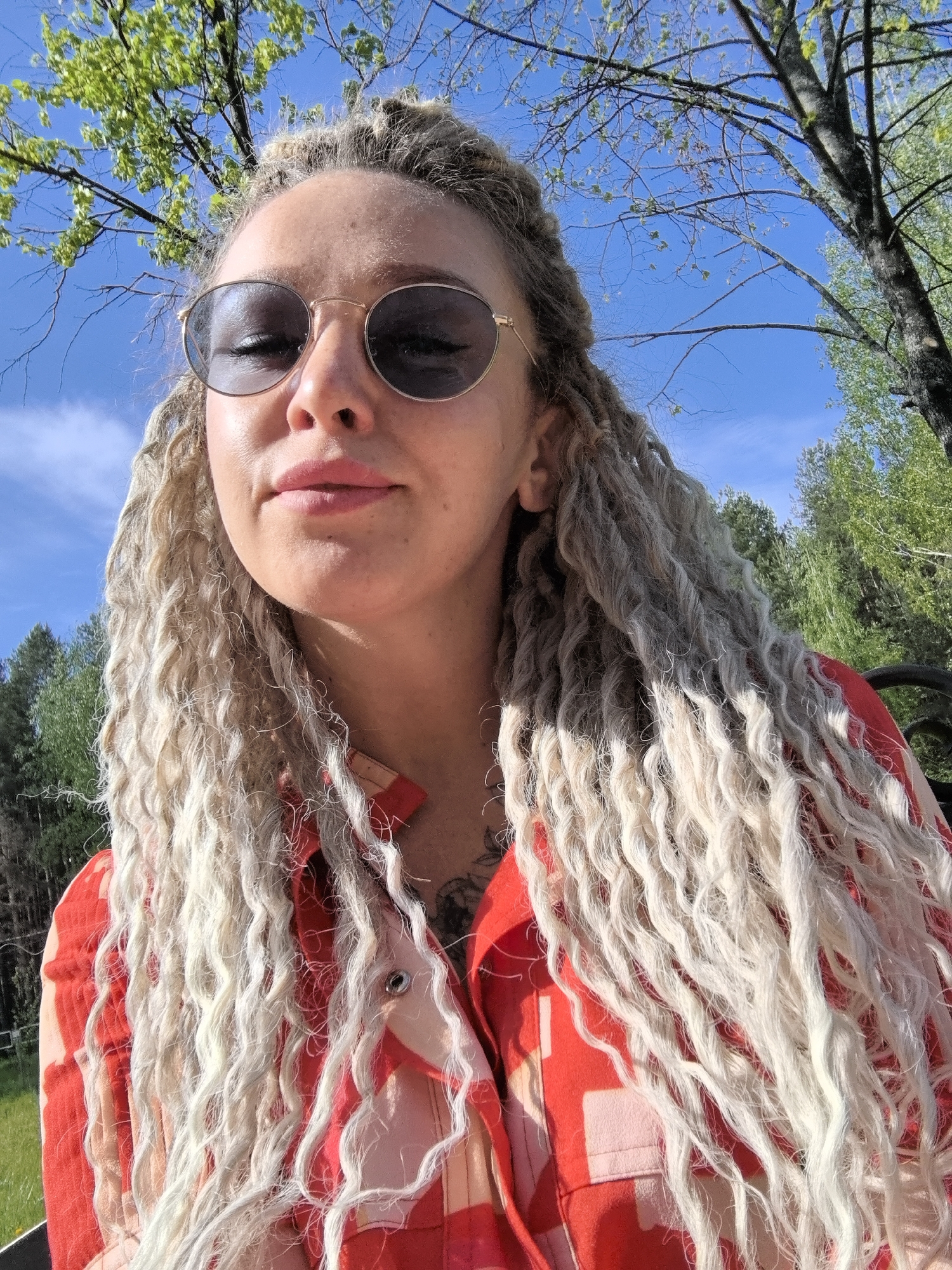A woman with blonde, curly dreadlocks is shown in a close-up, upward-angled shot. She is wearing round sunglasses with a dark tint. She has a red patterned shirt on. The background features a blue sky with a few clouds, as well as some green trees and foliage. It is impossible to determine the specific location in the city of Chaikovsky, Russia, based on the given information.