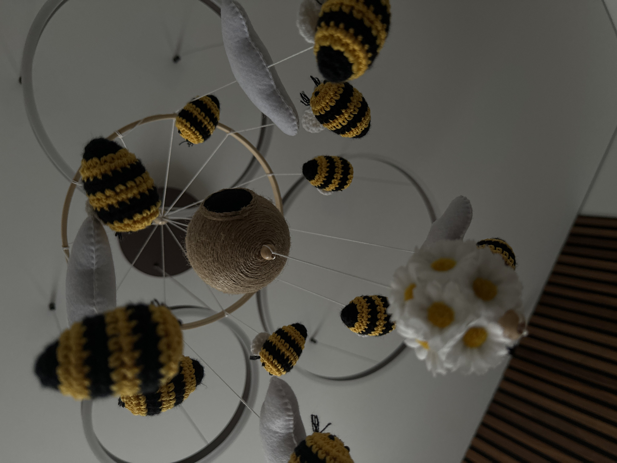 The image shows a hanging mobile with crocheted bees, white felt wings, a yarn-wrapped ball, and a white floral element. The mobile's frame is made of concentric metal and wooden rings. It is suspended from a light-colored ceiling. A portion of a wood-paneled wall is visible in the bottom right corner. The scene appears to be indoors, likely in a child's room. There is no indication of the location being Akulovo (Gp Kubinka), Russia.