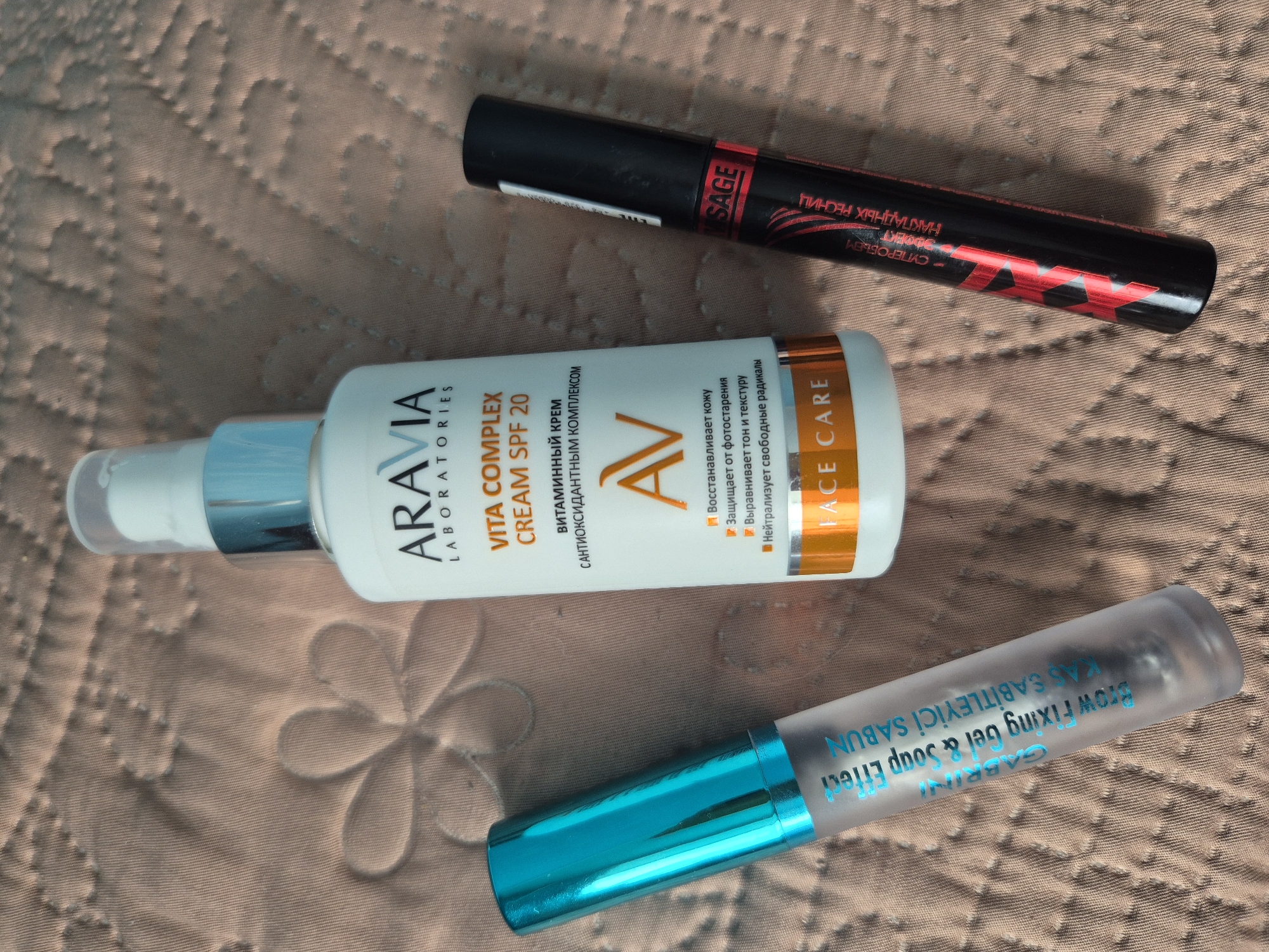The image displays a flat lay arrangement of three cosmetic items on a textured, light brown fabric surface, possibly a quilted blanket or tablecloth with a floral pattern. The items are positioned diagonally across the frame.
On the left is a translucent cylindrical tube with a metallic blue cap, likely a brow product, with text in blue that reads "GABRINI Brow Fixing Gel & Soup Effect KAS SABITLEYICI SABUN".
In the center is a white pump bottle with a blue collar and a clear cap, labeled "ARAVIA LABORATORIES VITA COMPLEX CREAM SPF 20". Below this, text in Russian indicates the product's benefits. The bottle's base has an orange band with the words "FACE CARE".
On the right is a black mascara tube with a prominent red and black label. The label features stylized red lettering that appears to spell "VISAGE" and other text in Russian, including "СУПЕРОВЫЕМ ЭФФЕКТ" and "НАКЛАДНЫХ РЕСНИЦ".
The lighting suggests an indoor setting, possibly a home. There are no people or animals visible in the image. The background fabric has a soft, dimpled texture, and the objects are centrally placed, with the camera positioned directly above. The image does not contain any visual information that would allow for the determination of its location within Kokshetau, Kazakhstan.