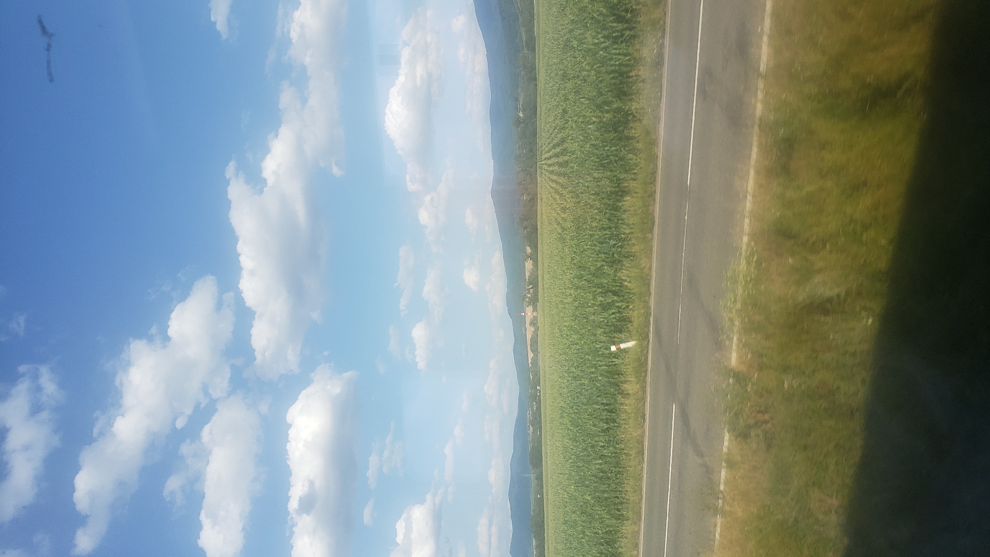 A wide shot captures a sunlit rural landscape under a bright blue sky dotted with fluffy white clouds. In the foreground, a paved road with a white line runs horizontally across the frame, bordered by overgrown green grass. Beyond the road, a vast, neatly cultivated field of tall green crops, likely corn, stretches into the distance. In the background, rolling hills and mountains form the horizon, with a few scattered buildings and trees visible. The scene evokes a sense of rural tranquility and agricultural abundance.
