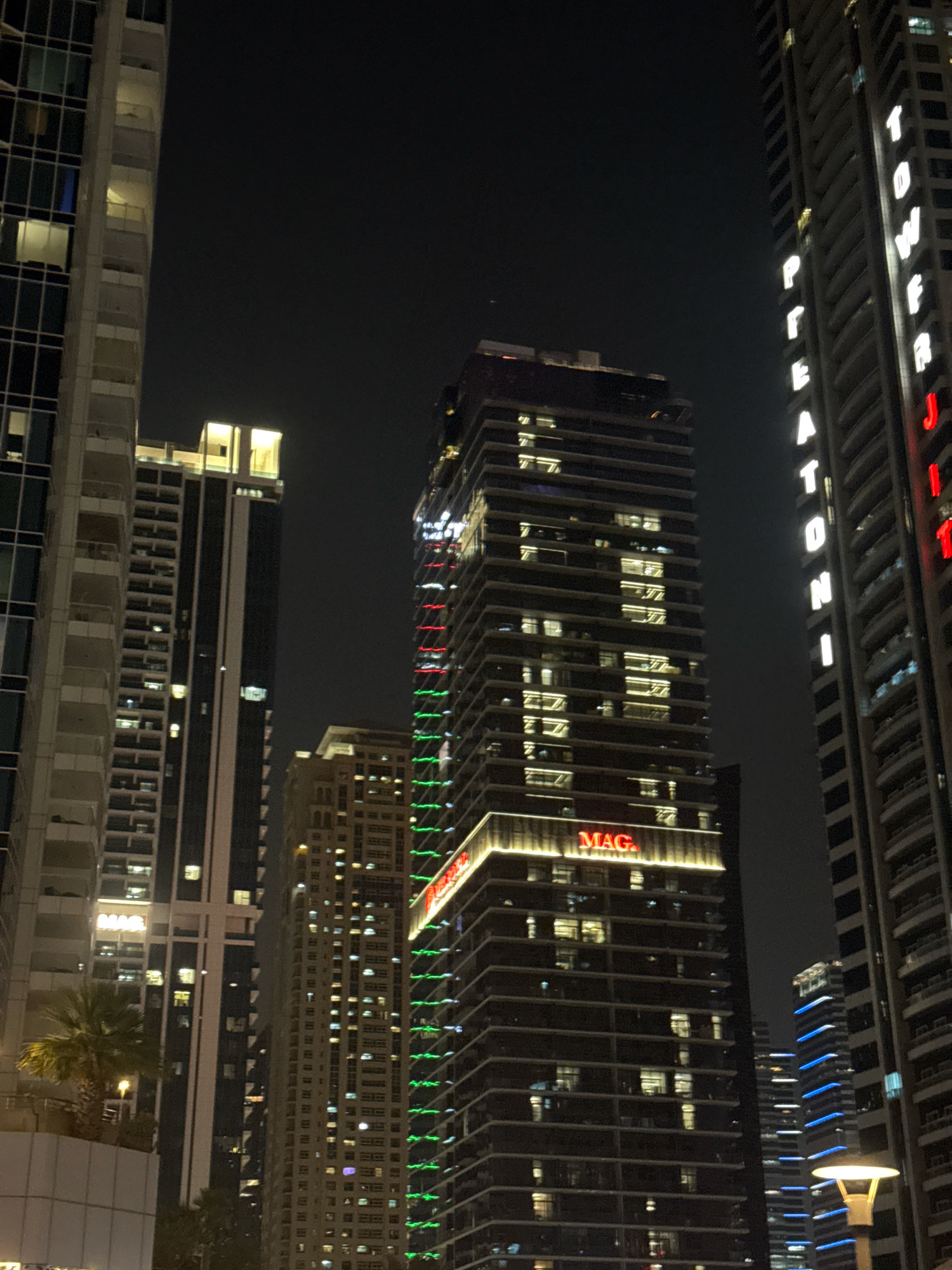 The image shows a nighttime view of several illuminated skyscrapers in Dubai, United Arab Emirates, likely in the Dubai Marina area given the density of tall buildings. The central focus is a very tall, dark skyscraper with many lit windows arranged in a grid pattern. At its base, a prominent sign reads "MAG." in red illuminated letters. Another skyscraper to the right, also very tall and dark, has vertical white illuminated letters spelling out "TOWER J" and partially visible letters of another word. This building also features red and green lights running vertically down its facade. A third, lighter-colored skyscraper is visible to the left, with a more classic architectural style and numerous balconies. In the background, several other buildings are visible, some with blue and white lighting. A street lamp with a white light is positioned in the foreground at the bottom right. A palm tree is visible on a lower level in the mid-left of the image. The overall scene depicts a modern urban landscape at night, characterized by extensive artificial lighting and towering architecture.