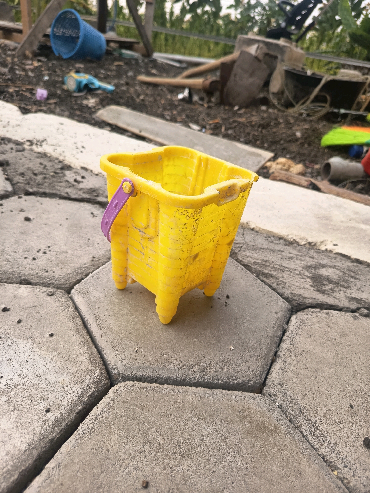 In the city of Sidorejo, Indonesia, a yellow plastic toy bucket with a purple handle sits on a paved surface made of interlocking hexagonal concrete blocks. The bucket is positioned in the center of the frame, with its legs resting on the blocks. In the background, various discarded items and debris are visible, including a blue plastic basket, a blue and yellow toy shovel, and what appears to be construction materials like wood and a wheelbarrow. The overall environment suggests an outdoor, possibly a yard or construction area, with greenery in the distance.