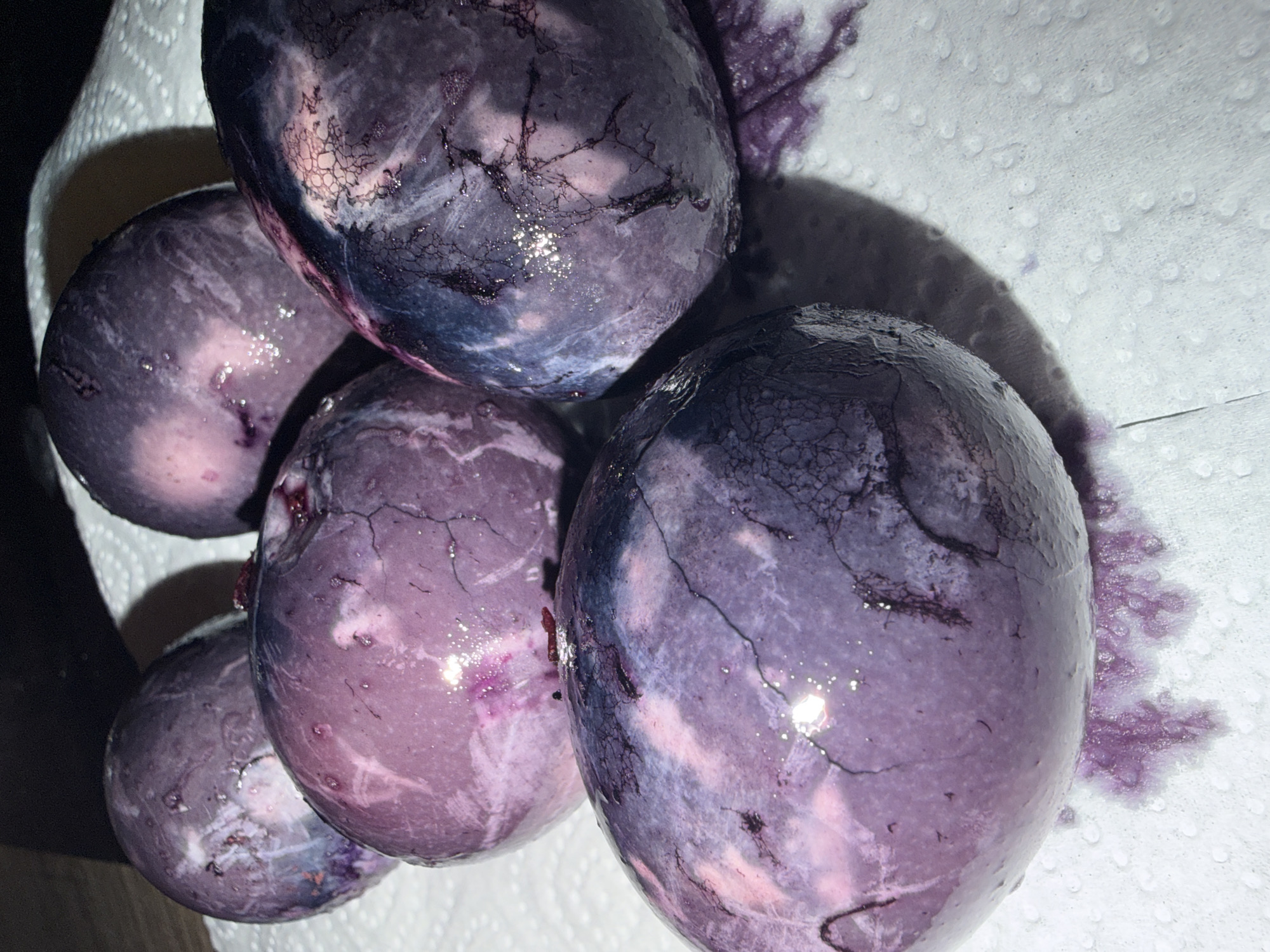 The image shows five purple-dyed Easter eggs resting on a white, textured surface, likely a paper towel. The eggs exhibit a marbled or cracked pattern, suggesting they were dyed using a technique involving cracked shells. Some of the purple dye has bled onto the paper towel. The eggs are shiny, possibly due to moisture or a coating. The setting appears to be indoors. There is no indication of this image being taken in or around Chaikovsky, Russia. There is no mention of any people or interactions. The scene consists of dyed Easter eggs on a paper towel.