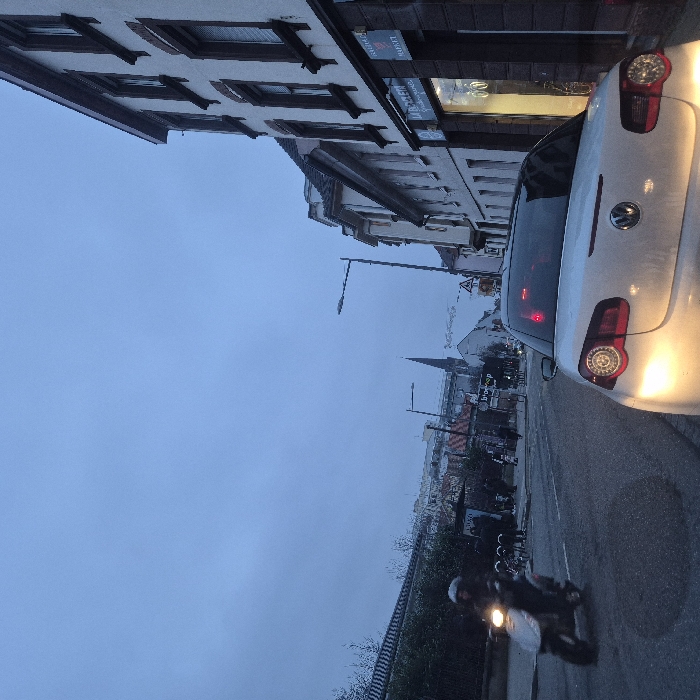 An urban street scene shows a white car with illuminated taillights on the right. A scooter with its headlight on drives on the same side. Buildings line the left side of the street, with visible storefronts. Street lights are placed intermittently. In the distance, a steeple rises above other buildings, suggesting a town or city view under a cloudy sky.