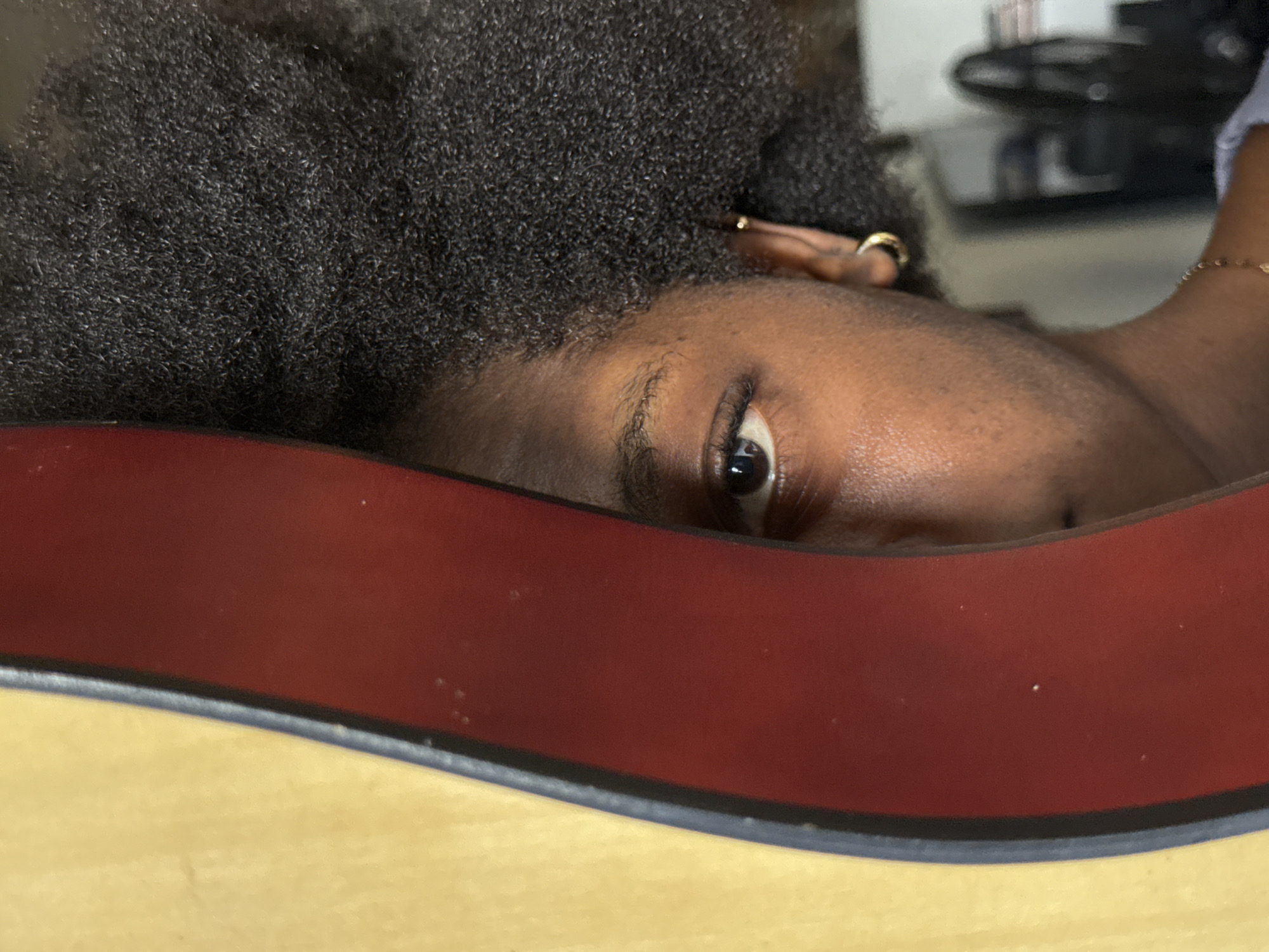 This is a close-up, self-portrait taken in Ado, Nigeria, featuring the right side of a person's face and head. The subject's dark, curly hair is prominently displayed. A portion of their right eye, with visible eyelashes and a brown iris, is visible. The person is wearing a gold hoop earring in their right ear and a delicate gold necklace around their neck. A curved, reddish-brown object is positioned in the foreground, partially obscuring the left side of the subject's face, suggesting the use of a mirror or a reflective surface. The background is blurred but hints at an indoor environment with some objects visible, including what appears to be a fan.