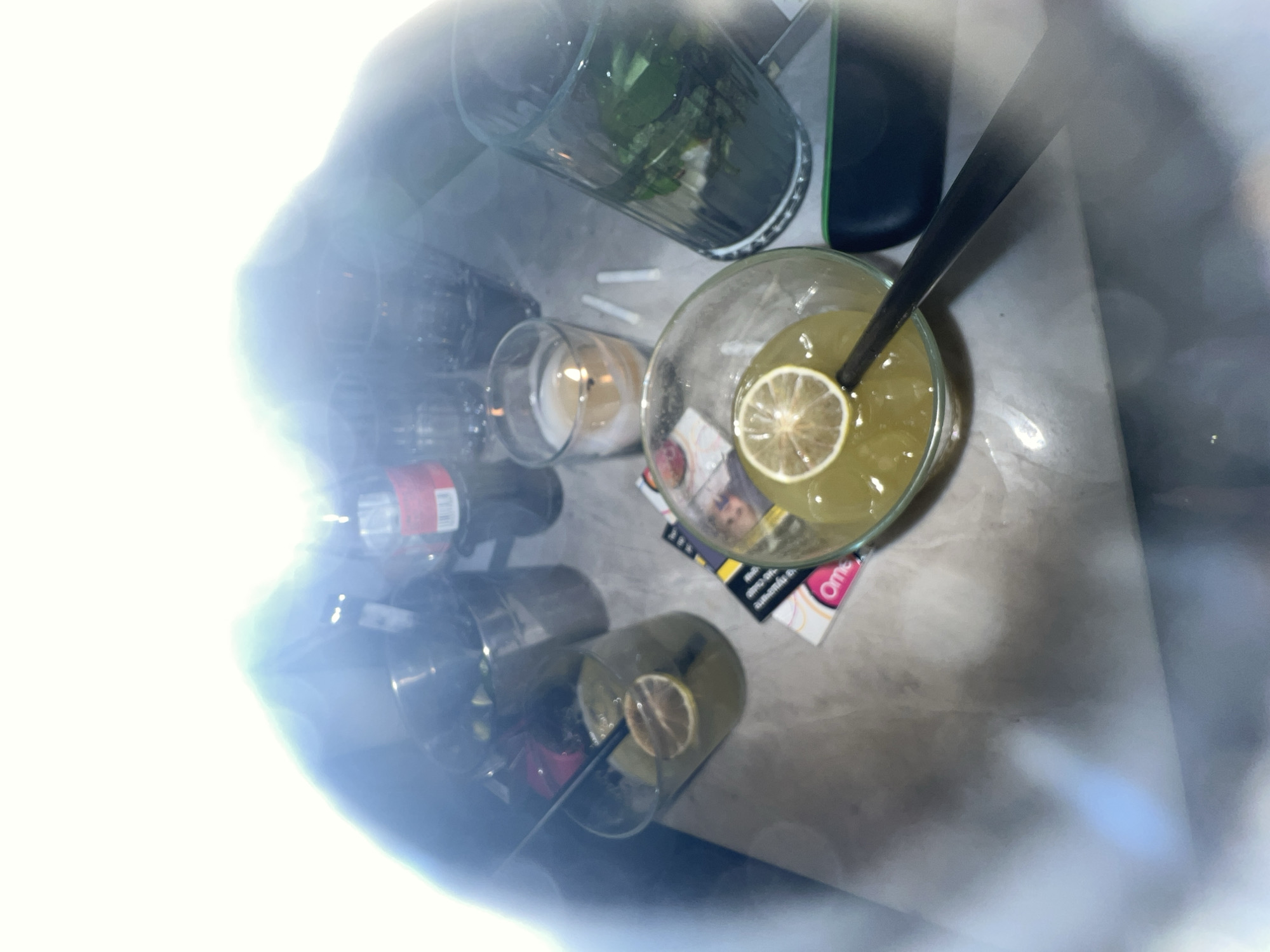 The image shows an overhead view of a table. On the table are two glasses of beverages with lemon slices and straws, a lit candle in a glass container, cigarettes, a vase with green plants, a bottle, and condoms. The location is potentially Plovdiv, Bulgaria, given the prompt's instruction. No people are visible.