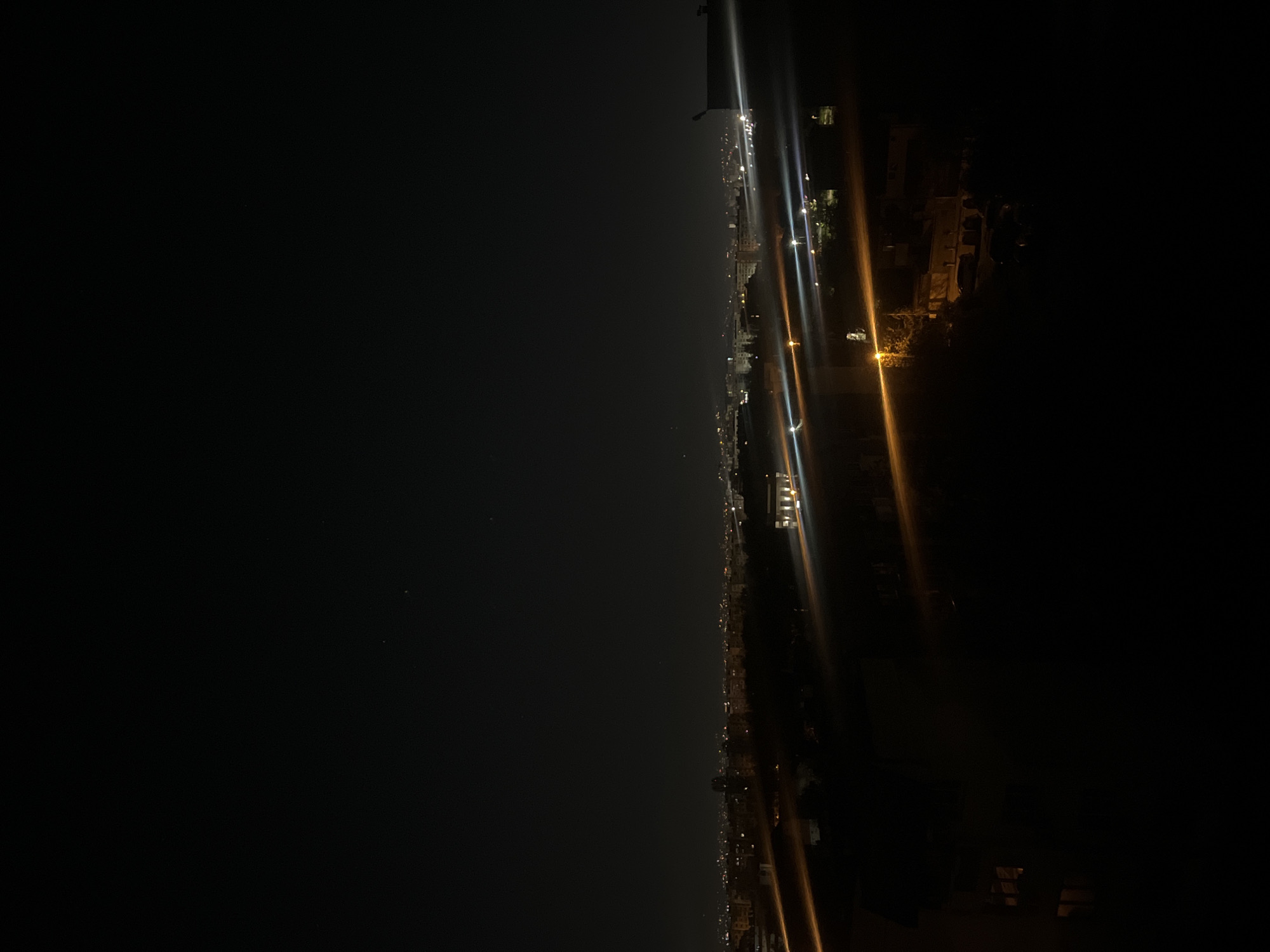 A panoramic night view of a sprawling city, likely Sofia, Bulgaria, is captured from a high vantage point. The scene depicts a vast urban landscape illuminated by countless artificial lights, stretching horizontally across the lower portion of the frame. The upper two-thirds of the image consist of a deep, dark night sky, suggesting a clear evening.
The city lights form a bright, continuous band, showcasing the density of the urban area. Brighter streetlights in the closer foreground and mid-ground create prominent vertical lens flares, varying in color from warm amber and orange to cooler white. Further into the distance, the lights become smaller and more numerous, blurring into a luminous haze as the city recedes towards the horizon. Outlines of buildings, rooftops, and other urban structures are dimly discernible amidst the glow. The extreme dark areas of the sky indicate minimal light pollution directly above, though the city's glow is evident below.
No specific human activity or event is visible; the image captures the static grandeur of an illuminated city at rest. The time of day is clearly night. The weather appears clear, with no clouds visible. The extensive urban sprawl and density of lights are consistent with a large city like Sofia. No text is visible in the image.