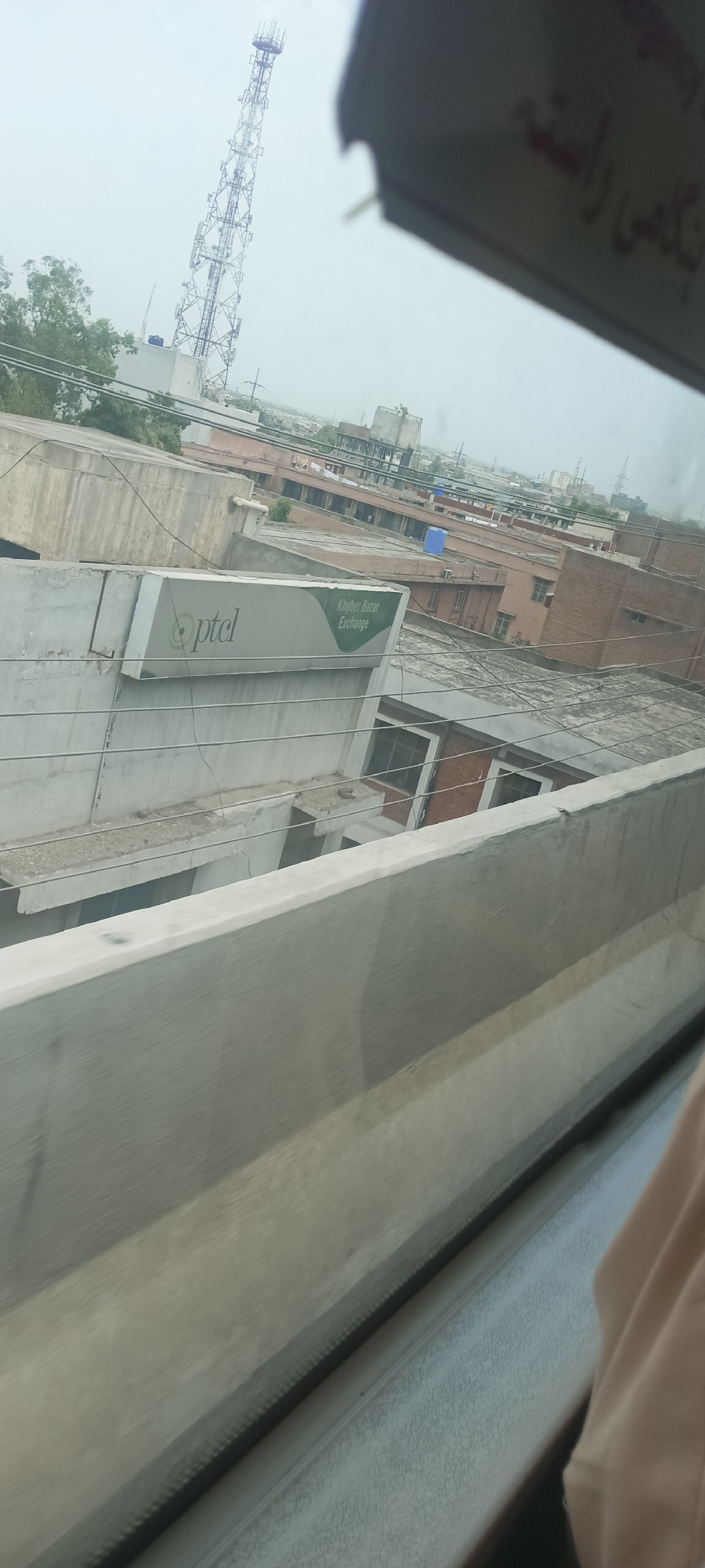 The image shows a view from a moving vehicle in Alam Khan Kalay, Pakistan. In the background, a tall cell tower stands against a cloudy sky. Below the tower, a cluster of buildings with flat roofs and brick facades are visible. One building has a prominent sign that reads "ptcl Khyber Bazar Exchange". In the foreground, a concrete railing suggests the perspective is from a bridge or elevated structure. A person's arm, wearing a light-colored garment, is partially visible on the right side of the frame, indicating the presence of an occupant within the vehicle. The overall scene depicts an urban or semi-urban environment with infrastructure and commercial activity.
