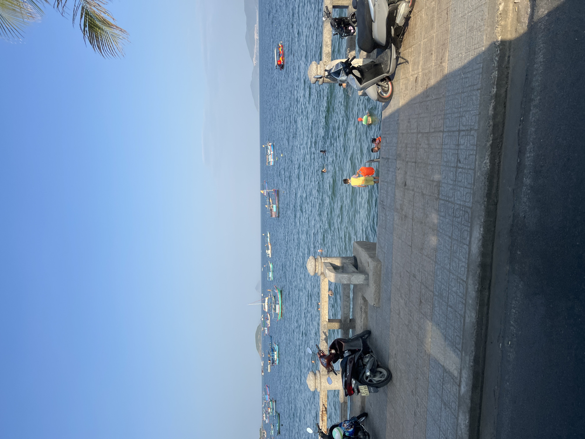 This image captures a picturesque scene at the beach in Nha Trang, Vietnam, likely along the main beachfront promenade. The clear blue sky meets the calm, azure sea, dotted with numerous small fishing boats. A few people are enjoying the water, some wading in the shallows, others swimming further out. On the paved walkway overlooking the sea, several motorbikes are parked, indicating a common mode of transportation for visitors and locals. The walkway itself is lined with decorative stone pillars and a low barrier, suggesting it's a popular spot for strolling and enjoying the ocean views. The presence of palm fronds from a nearby tree on the right adds to the tropical ambiance of the location. The overall atmosphere is one of leisure and relaxation, characteristic of a seaside resort town.