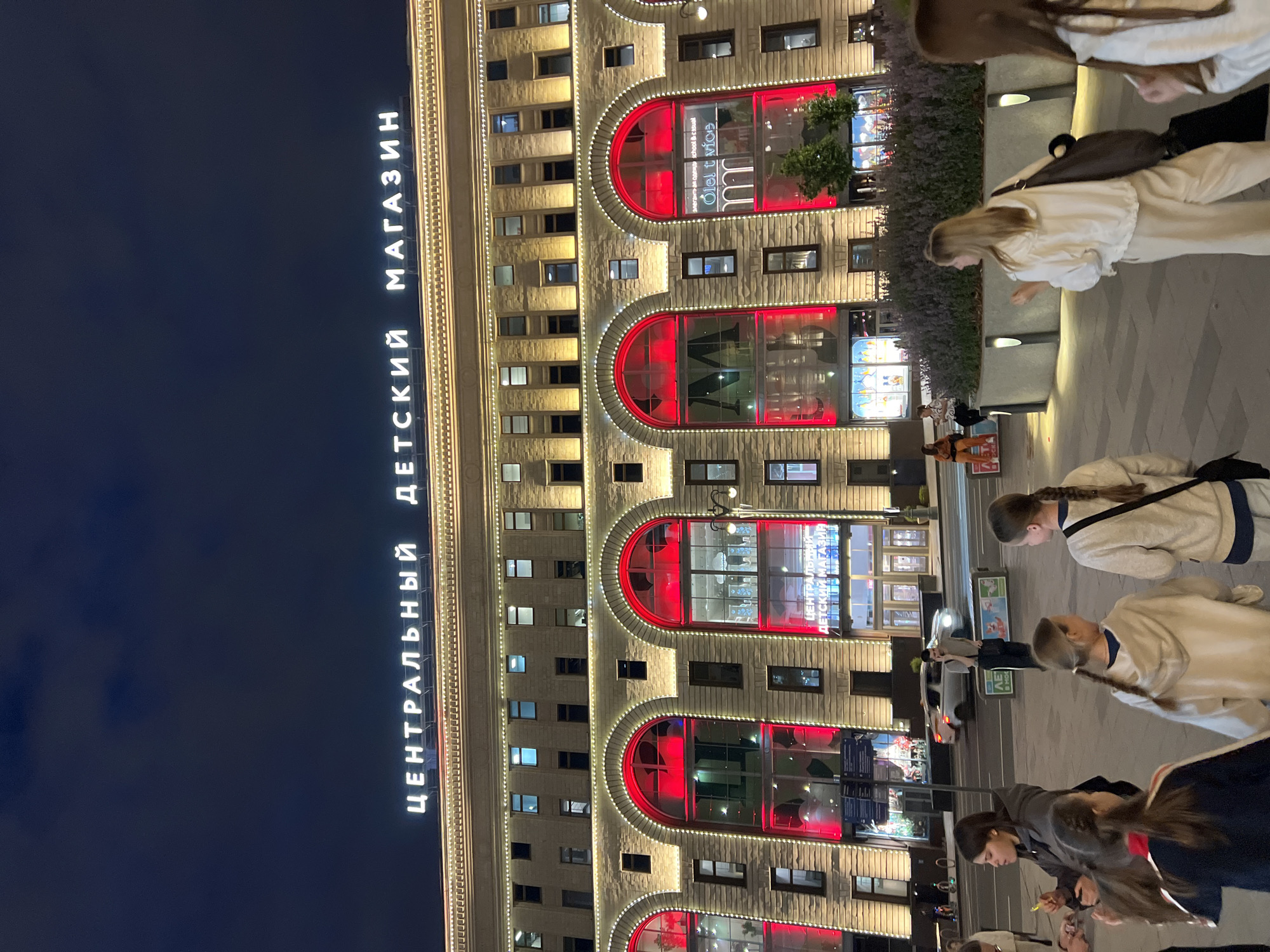 The image shows the Central Children's Store (TsUM) in Moscow, Russia, at night. The iconic building, illuminated with warm yellow lights along its facade and bright red lights within its arched windows, is a prominent feature. The Cyrillic inscription "ЦЕНТРАЛЬНЫЙ ДЕТСКИЙ МАГАЗИН" (Central Children's Store) is visible in large white letters across the top of the building.
In the foreground, several individuals, appearing to be families and friends, are walking on a paved public area. Some are walking towards the camera, while others are walking away. Among them are at least four young girls, some with their hair in braids, and two adult women. One woman is speaking on a mobile phone while standing in the middle of the street, with cars passing behind her. A silver car is visible on the road.
Behind the people and the street, the Central Children's Store stands majestically, its impressive architecture clearly visible. The scene captures a lively evening atmosphere in a well-known Moscow landmark.