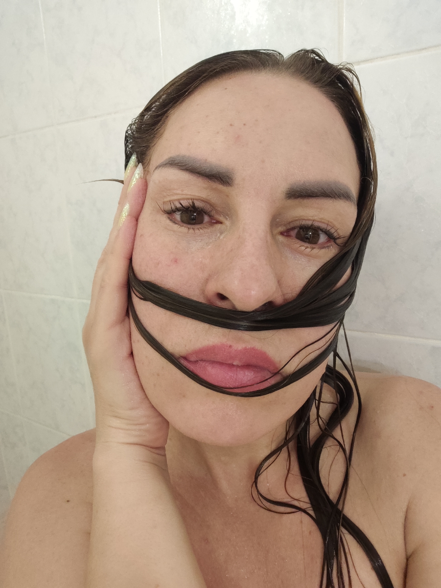 A close-up, waist-up, indoor, point-of-view photograph shows a woman in what appears to be a shower. The woman is facing the camera, her right hand is on the left side of her face, and her dark, wet hair is draped across her mouth and chin. Her left eye is visible, and she is wearing subtle makeup. The background consists of a tiled wall, common in a shower or bathroom setting. The lighting is diffused, suggesting an interior light source. The scene does not contain any specific identifying landmarks or features that would indicate a location within Almaty, Kazakhstan.