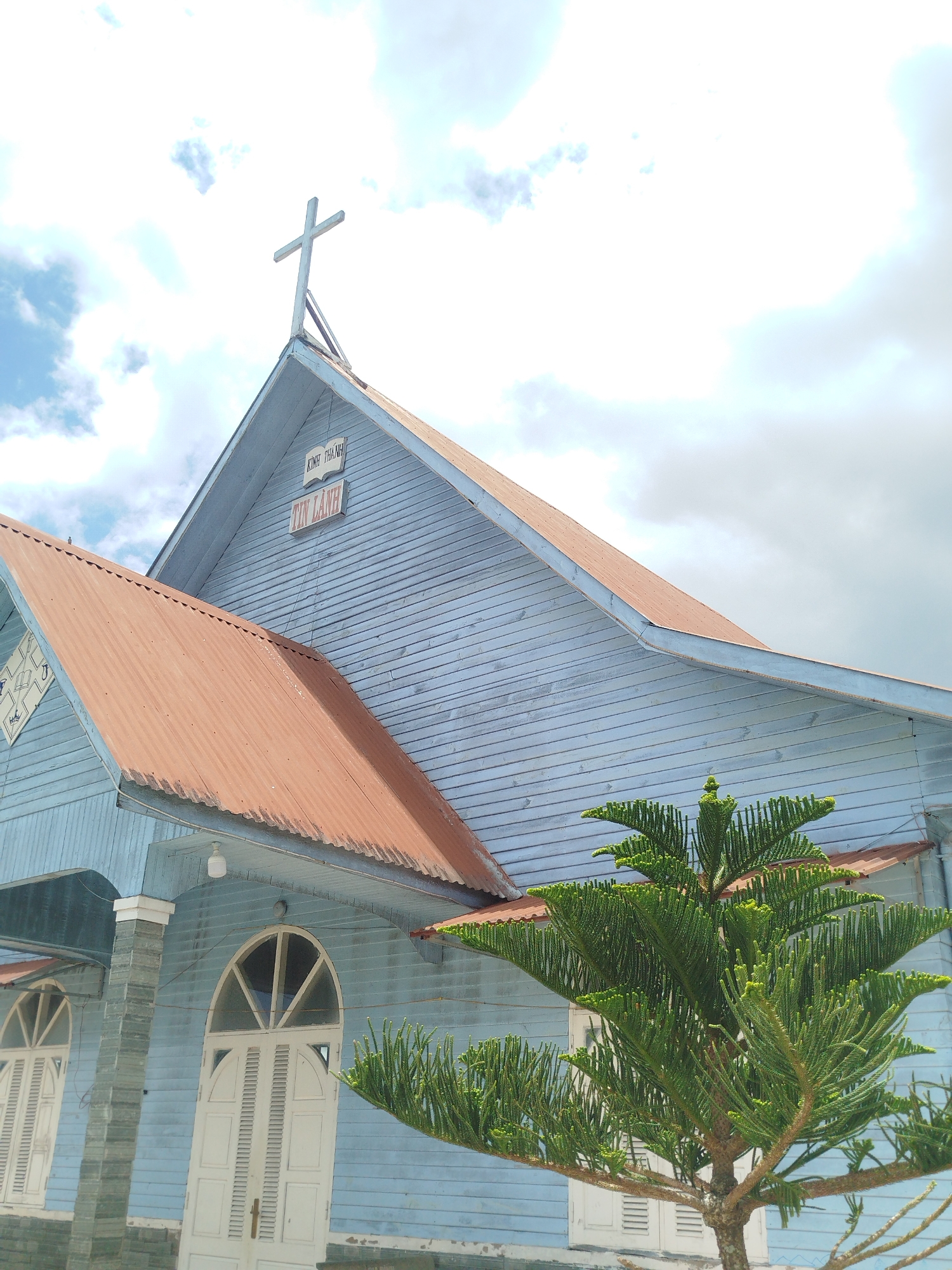 This is a low-angle, close-up shot of a light blue wooden church in Thôn 6, Vietnam. A prominent red metal roof juts out diagonally from the left side of the frame, overlapping a smaller section of the church's light blue wall. The church's facade is constructed of horizontal wooden planks painted light blue, with white trim and columns. Two arched white doors with intricate wooden detailing are visible on the lower portion of the building. Above the doors, there are semicircular windows. The roof of the church is topped with a white cross. To the right, a green evergreen tree with dense, spiky foliage partially obscures the church. The sky above is a mix of bright white clouds and patches of blue. A sign on the church wall reads "KINH THANH" and "TIN LANH".