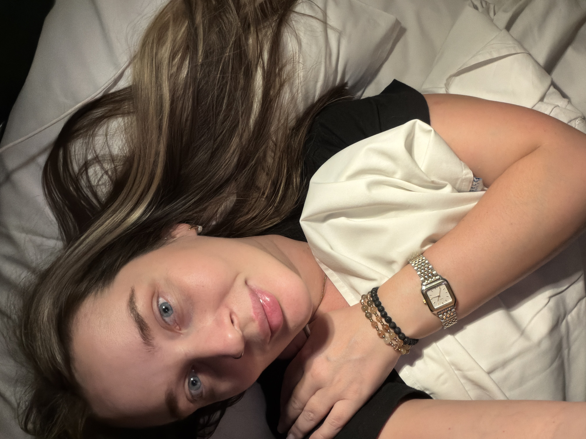 A woman is lying in a bed with white sheets and pillows. She is wearing a black t-shirt and has long brown hair with blonde highlights. She has blue eyes, fair skin, and a nose piercing. On her wrist, she is wearing a silver watch and beaded bracelets. The lighting is soft and diffused. There is no indication of the city of Moscow, Russia, or any external location.