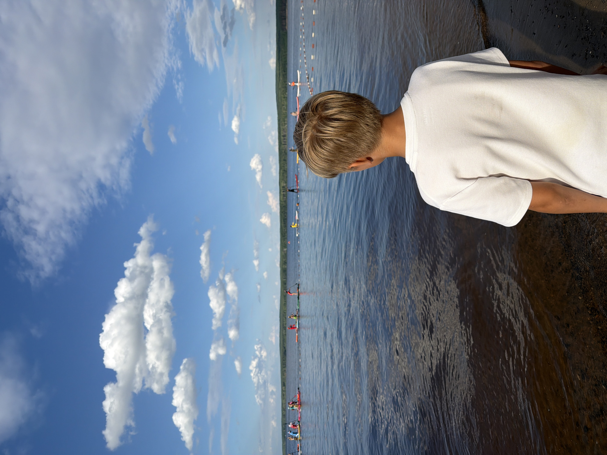 The image depicts a young boy with blonde hair, wearing a white waffle-knit t-shirt, standing on the shore and looking out at a body of water. The water is calm and reflects the blue sky with scattered white clouds. In the distance, several people are visible on paddleboards, participating in an activity, possibly a race, indicated by a string of buoys in the water. The shoreline is composed of sand and small pebbles. The background features a line of trees. The scene appears to be set on a sunny day. This scene is consistent with locations along the Kama River in Chaikovsky, Russia, which is known for its recreational water activities.