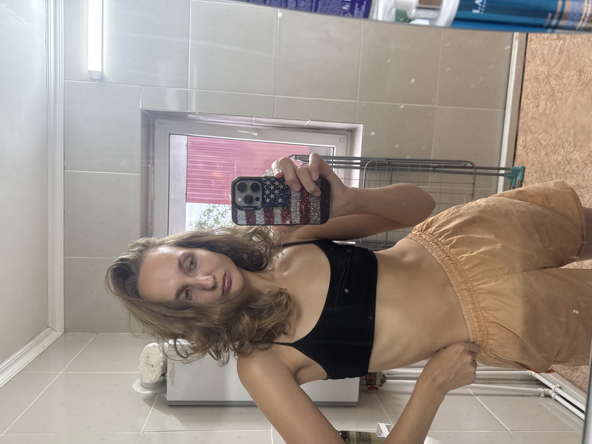 A young woman with medium-length, wavy brown hair takes a mirror selfie in a bathroom. She is wearing a black spaghetti-strap bra and orange athletic shorts. Her left hand is placed on her hip, while her right hand holds a smartphone with an American flag-patterned case. The bathroom features beige tiled walls, a white tiled ceiling with a fluorescent light fixture, and a window with a red-patterned curtain visible through it. A white cabinet or storage unit is to the left of the woman, and a drying rack is visible behind her. A bottle of what appears to be skincare or cleaning product is partially visible on the right side of the frame, reflecting in the mirror. The flooring is a textured, reddish-brown material.