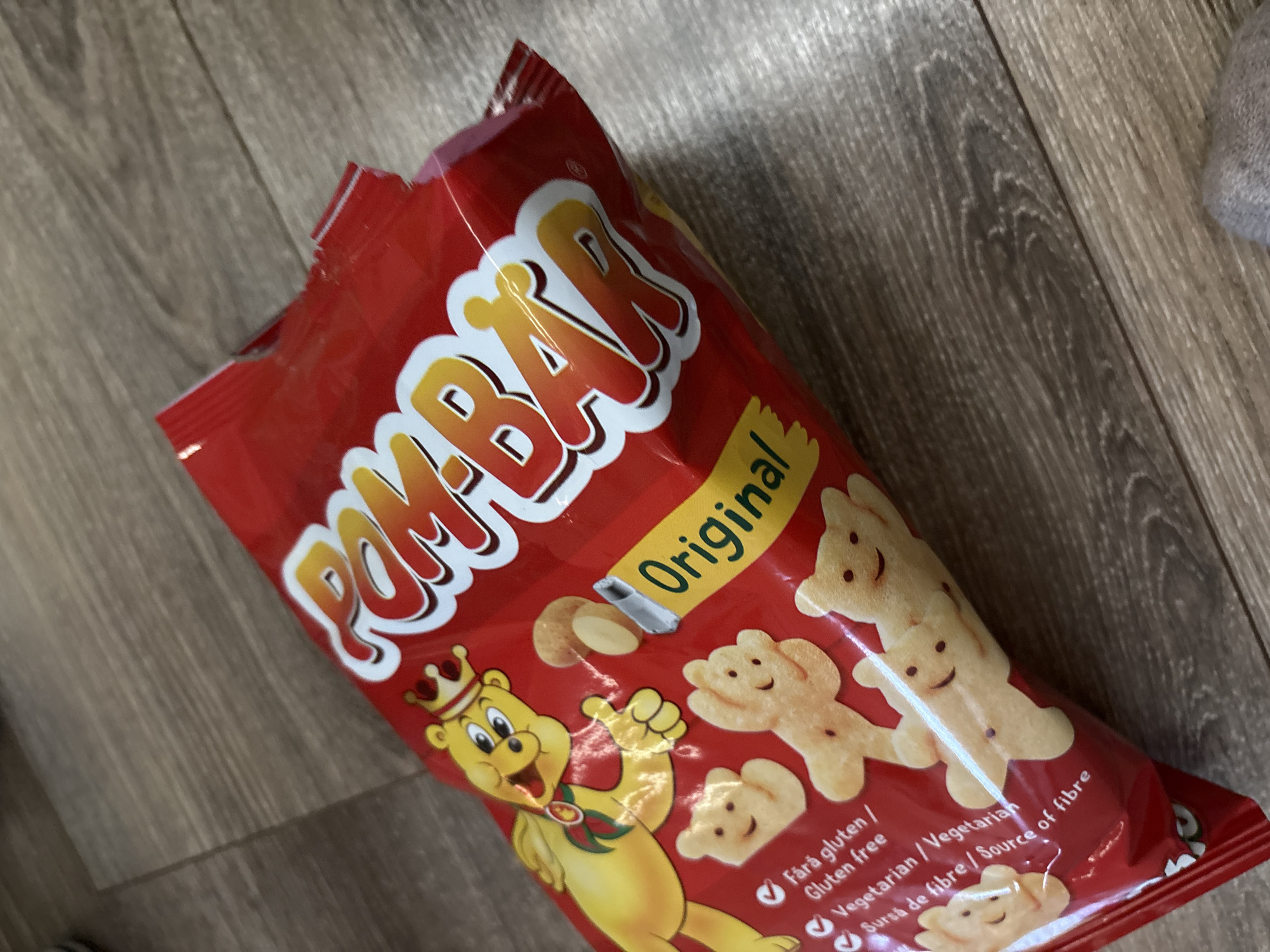 A slightly open bag of "Pom-Bär Original" potato chips sits on a light brown wood-grained surface. The bag is predominantly red, with the product name "POM-BÄR" in large, stylized white letters outlined in yellow. A yellow banner with "Original" written in black diagonally is visible. The bag's design features cartoon bear-shaped chips, and a cartoon bear character wearing a crown. Text indicating "Fără gluten / Gluten free," "Vegetarian / Vegetarian," and "Sursă de fibre / Source of fibre" is printed with checkmarks in front. No people are visible, and the environment appears to be indoors. There is no indication of Burgas, Bulgaria in the image.