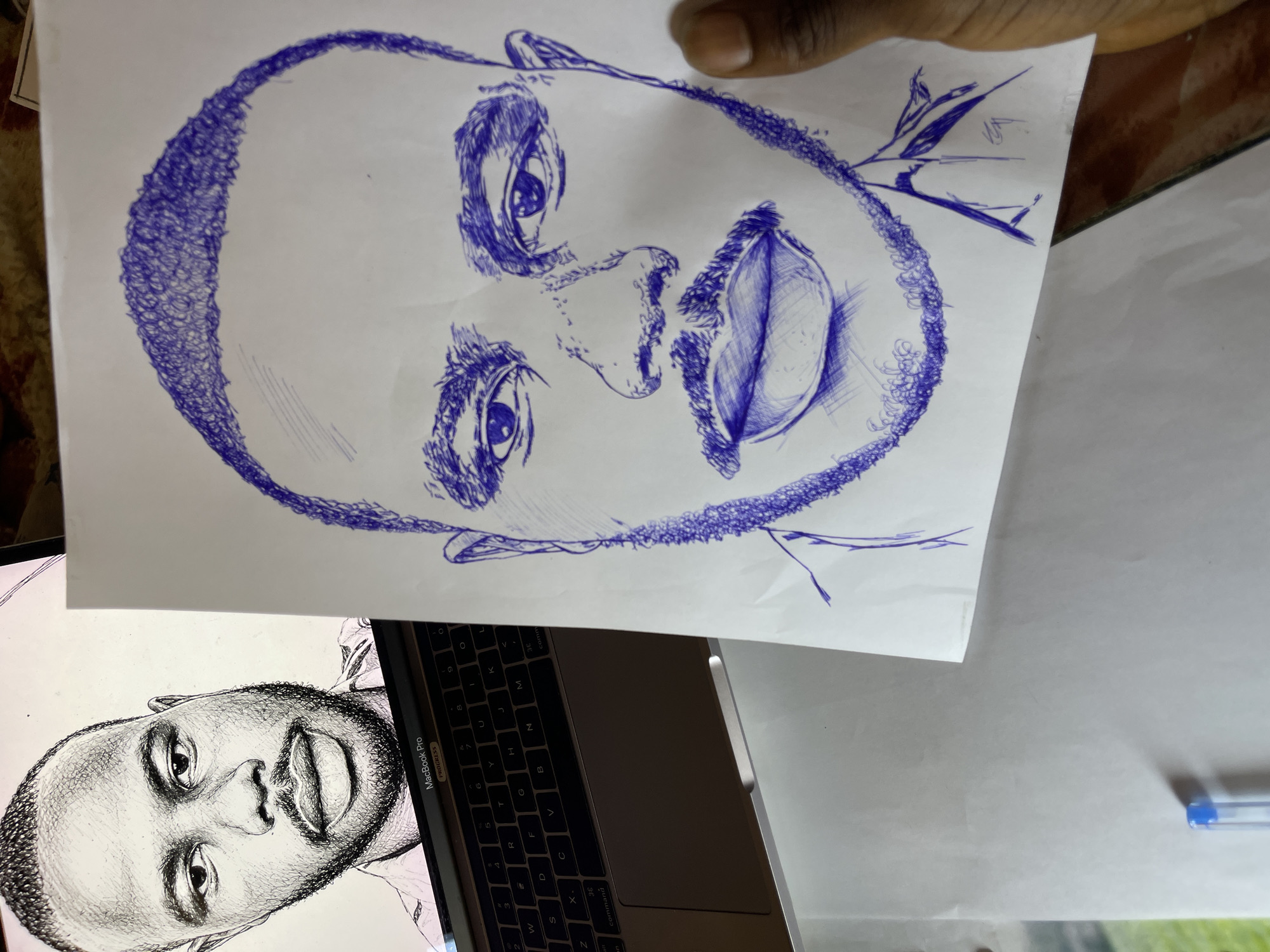 A sketch of a man's face drawn in blue ink on white paper is placed on a table. The sketch is a close-up, showing the man's eyes, nose, mouth, and beard. The man's head is bald with a short haircut. The sketch is detailed, with shading used to create depth and form. The paper is held by a person's hand, with a thumb visible on the right side of the frame.
To the left of the sketch, a laptop is open, displaying a portrait of the same man, also drawn in ink. The laptop screen shows the MacBook Pro logo. The keyboard of the laptop is visible, with keys labeled in English.
The background is blurred, but appears to be an indoor setting. A blue pen is visible at the bottom left of the frame, lying on a white surface.
The image does not provide any information about the specific location in the city of Jos, Nigeria.