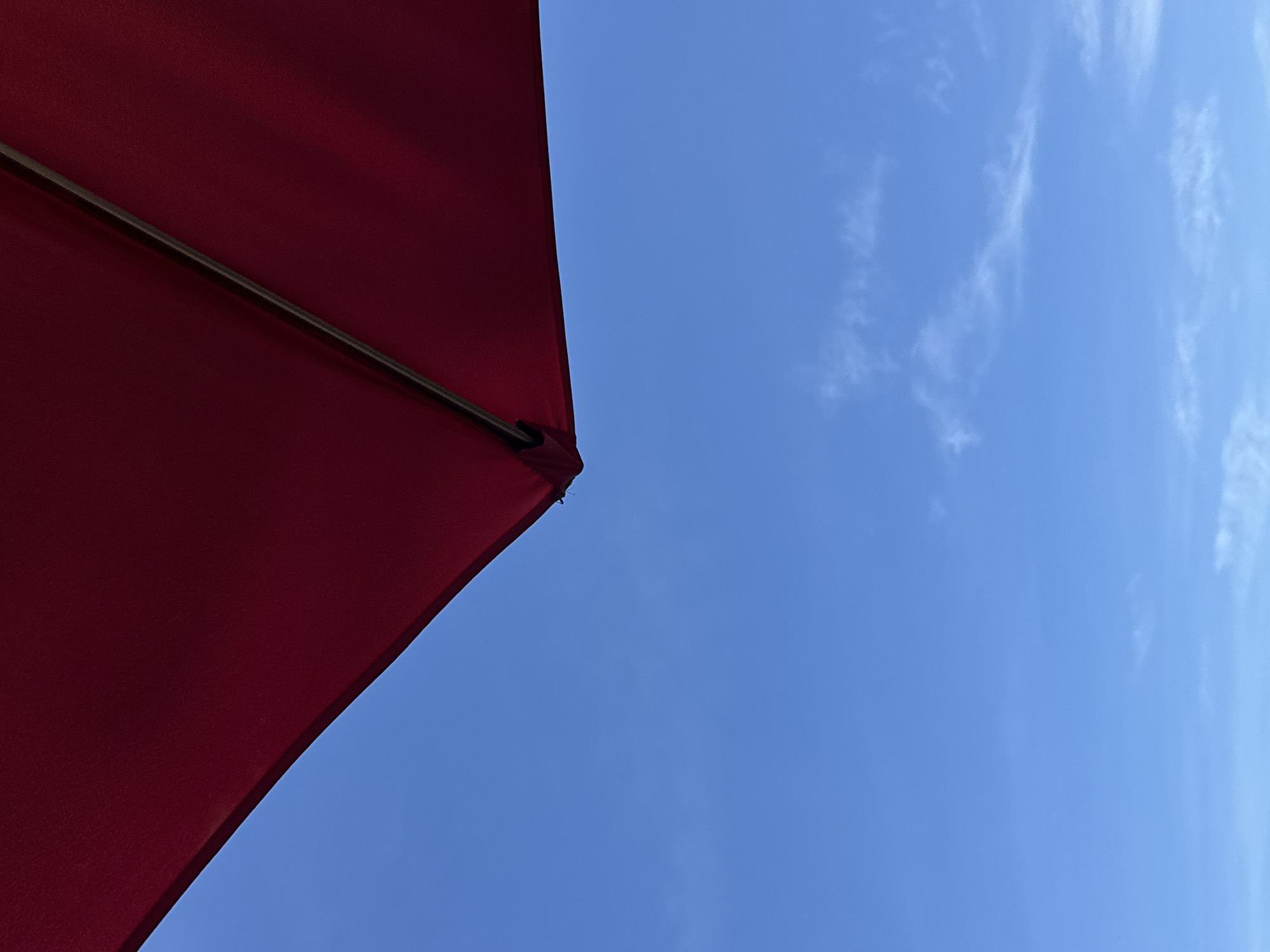 This is a close-up, low-angle outdoor shot of a red umbrella against a clear blue sky. The umbrella takes up the top portion of the frame, with its edge and a supporting metal rod clearly visible. The sky occupies the bottom two-thirds of the image and is a bright, cloudless blue, with only a few wispy, faint white clouds present. The image has a minimalist aesthetic, focusing on the contrast between the deep red of the umbrella and the expansive blue of the sky. There are no people or other objects visible in the frame. The context of Da Nang, Vietnam, is not visually evident within the image itself.