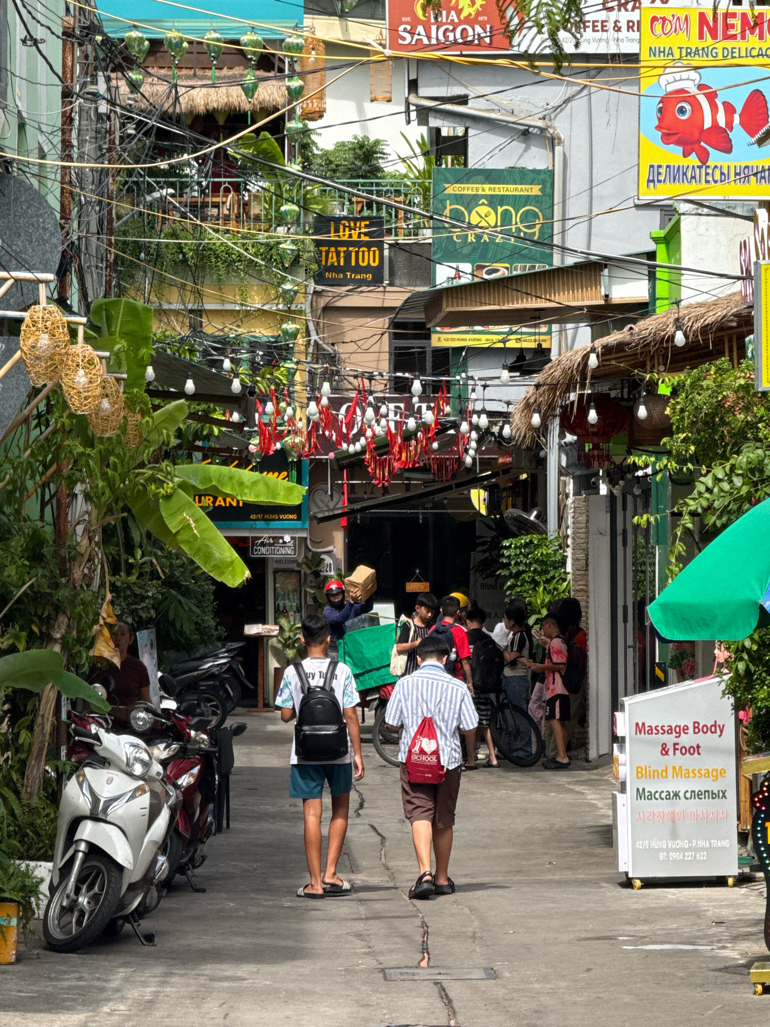 The image depicts a bustling street scene in Nha Trang, Vietnam. A narrow alleyway is lined with shops and businesses, many adorned with colorful signs and awnings. Overhead, a dense network of electrical wires crisscrosses the sky, a common sight in Vietnamese cities. In the foreground, two young men walk away from the camera, one wearing a striped shirt and shorts, the other in a white t-shirt and dark shorts, both carrying backpacks. Motorcycles are parked along the side of the street, and a white scooter is prominently positioned on the left. Several people can be seen gathered near shop entrances, engaging in conversations or using their phones. Various signs in Vietnamese and other languages advertise services such as "Coffee & Restaurant," "LOVE TATTOO," and "Massage Body & Foot." Red and white decorative elements hang from above, possibly indicating a festive occasion. The overall atmosphere is one of lively urban commerce.