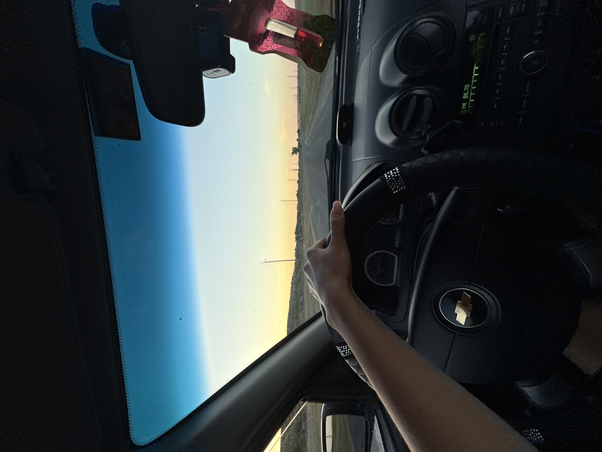 The image shows the interior of a car driving on a road at sunset. A person's left hand is on the steering wheel. The dashboard is visible, including the car's radio which is tuned to FM 88.70. A car freshener shaped like a gun is hanging from the rearview mirror. The exterior view through the windshield shows a road stretching into the distance, with utility poles on either side. The sky is clear with a gradient of blue and orange from sunset. There is no indication of the location being in Cioc-Maidan, Moldova.