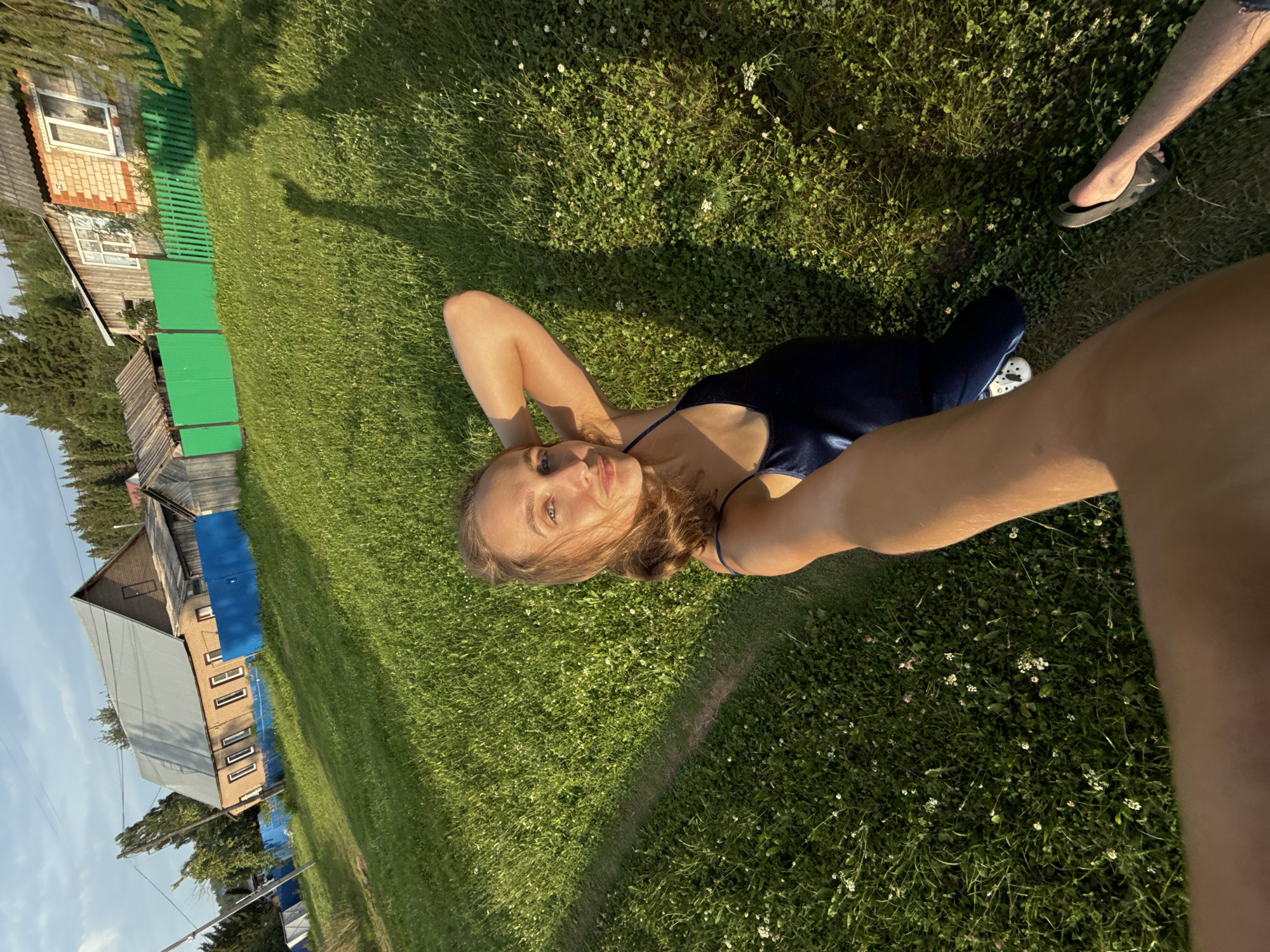 In Sharkan, Russia, a woman takes a selfie outdoors on a sunny day. She is lying on a grassy field with small white flowers scattered throughout. The woman is wearing a dark blue slip dress and has her right arm behind her head. In the background, there are houses and fences, one of which is painted bright green. Sunlight casts shadows on the grass.