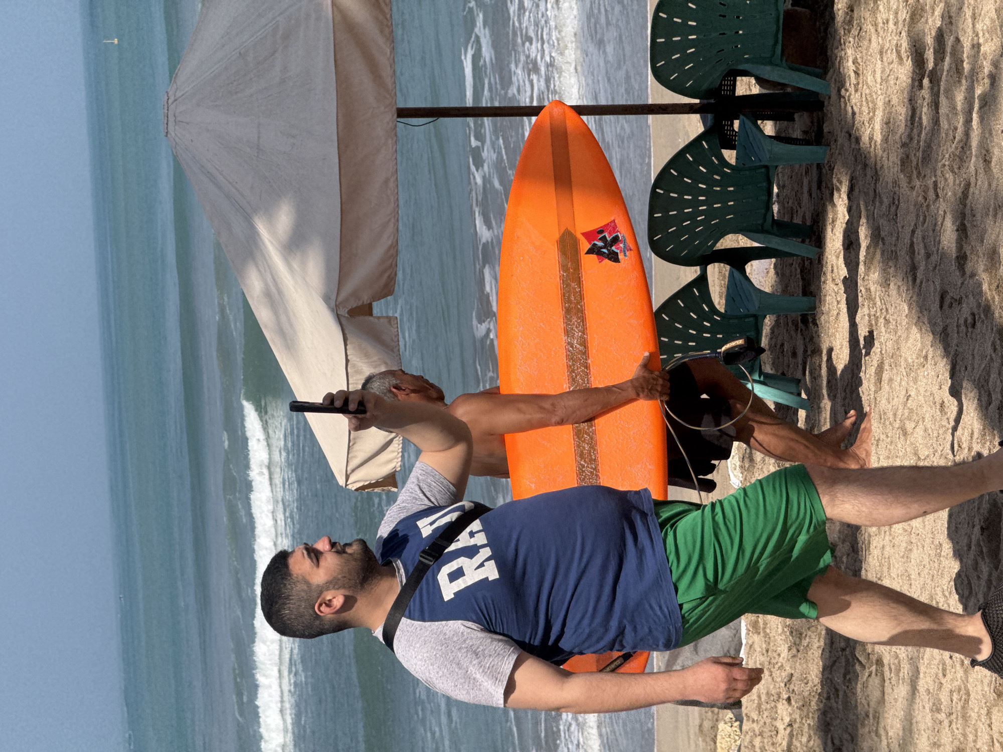 Two men are walking on a sandy beach in Kuta, Indonesia, with the ocean and waves in the background. The man in the foreground is wearing a navy blue t-shirt with white lettering that appears to say "RA", green shorts, and black shoes. He is holding a smartphone in his right hand and taking a selfie. He has a black strap across his shoulder. The second man, in the background, is shirtless and carrying a bright orange surfboard. Behind them are several green plastic chairs and a large tan beach umbrella. The sky is clear and blue, and the sun is shining. The scene suggests a typical day at a popular tourist destination.