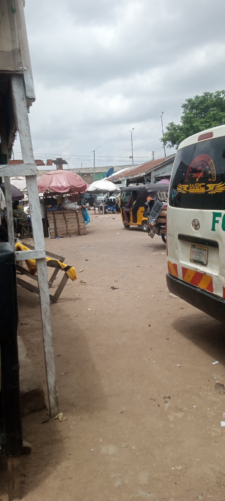 The image depicts a busy outdoor market scene in Jalingo, Nigeria, under a cloudy sky, suggesting it's likely daytime. The foreground features wooden market stalls with awnings providing shade. One stall has a collection of goods displayed under a pink umbrella, and another features sacks of what appears to be rice. A wooden bench with yellow fabric draped over it is also visible.
In the mid-ground, several vehicles are present, including a yellow auto-rickshaw with a driver and passenger. Behind it, a white van with red and yellow reflective markings on its rear is parked. The van has a sticker on the back window featuring a speedometer and text related to a rally championship, specifically "WRC 250 RALLY CHAMPIONSHIP". Visible text on the van's license plate reads "TARABA" and "MKD 553XA". Further into the scene, more market stalls and vehicles are discernible, along with some people seated at tables, suggesting a place to eat or rest.
The ground is unpaved and dusty, typical of many market environments in the region. Several streetlights are present in the background, indicating a developed urban area. The overall atmosphere appears lively and active, characteristic of a marketplace where commerce and daily life converge. The cloudy weather might suggest a humid or potentially rainy day, but the scene is clearly active.