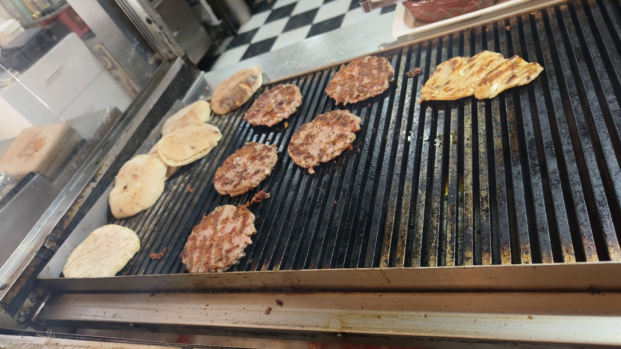 The image depicts a close-up, high-angle view of a commercial grill in what appears to be a food establishment in Предејане (варош), Serbia. The grill is a black, ribbed metal surface with several food items cooking on it. On the left side of the grill, there are several pieces of flatbread, some of which appear to be pitas, being warmed or lightly toasted. To the right of the flatbread, there are numerous oval-shaped, seasoned meat patties, likely burgers or ćevapi, in various stages of cooking, with visible grill marks and browning. In the upper right corner of the grill, a single piece of grilled chicken or pork is visible, also with prominent grill marks. The background of the scene includes a checkered floor pattern and what looks like a stainless steel kitchen counter or display area on the far left. The overall environment suggests a busy food preparation area.