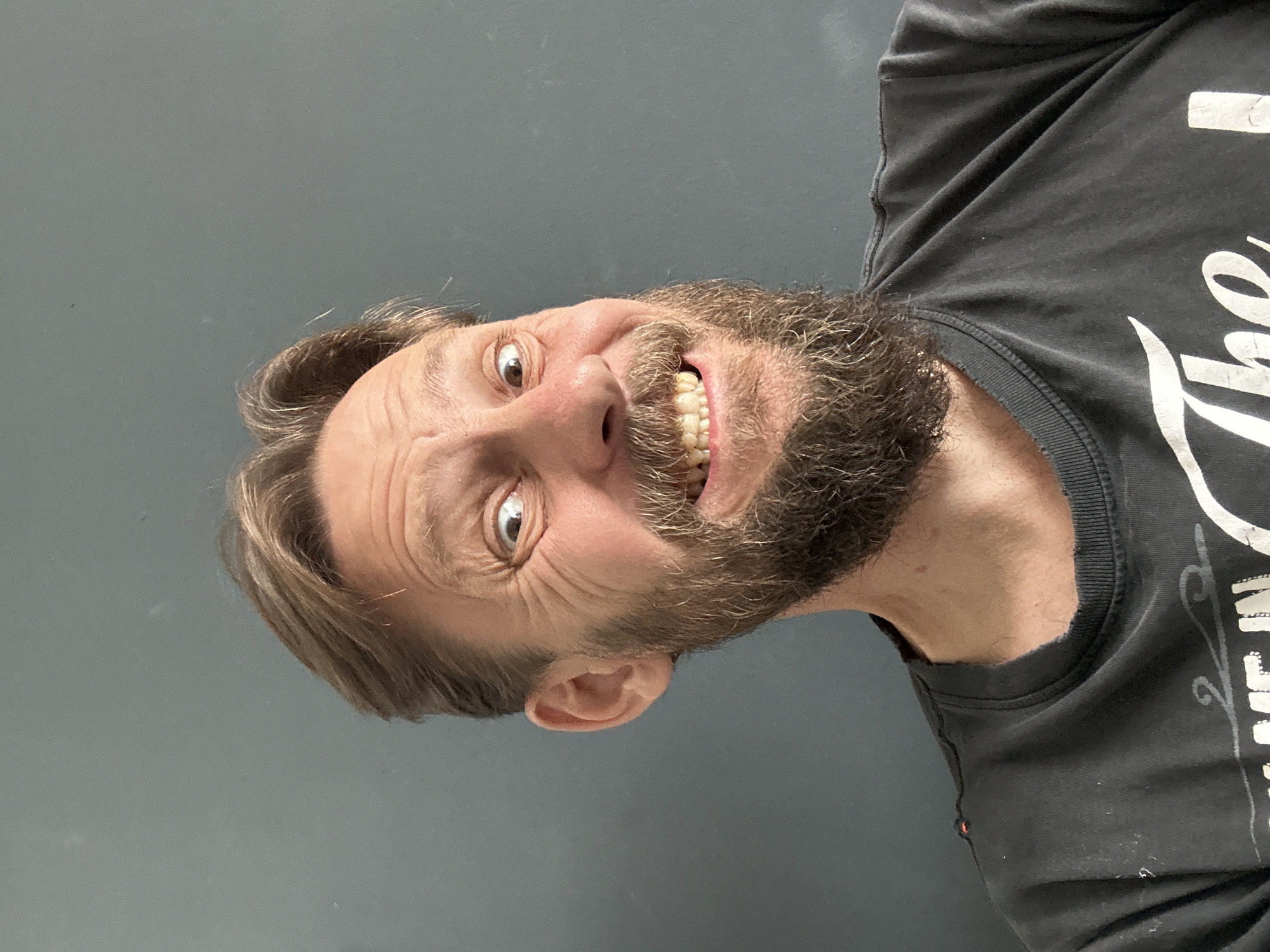 A man with light brown hair and a full, graying beard is grinning and looking directly at the camera. He is wearing a black t-shirt with white text. The background is a plain gray wall. The image does not show any details that would identify the location as Fethiye, Turkey.