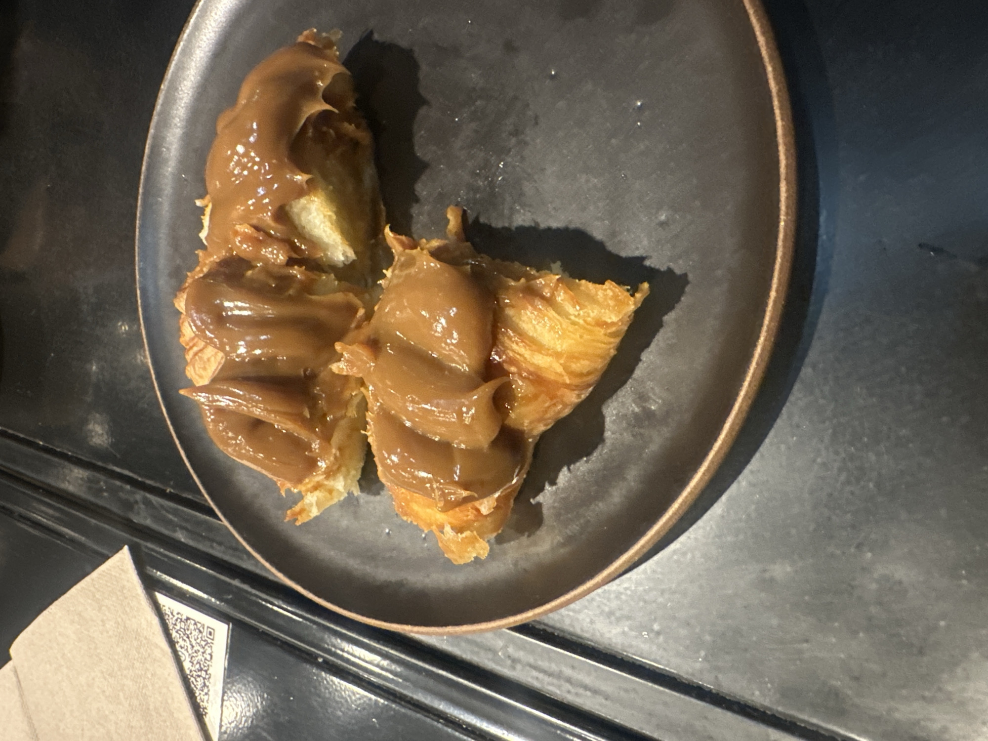On a dark gray plate with a brown rim sit two pieces of croissant topped with a thick, brown sauce. The plate rests on a dark surface, possibly a table, next to a folded beige napkin with a visible QR code. The photograph appears to have been taken indoors under dim lighting. The croissants are flaky and golden brown, contrasting with the smooth, glossy sauce that covers them. The overall setting is simple and suggests a dining experience, likely at a cafe or restaurant. The location is Unknown, Unknown.