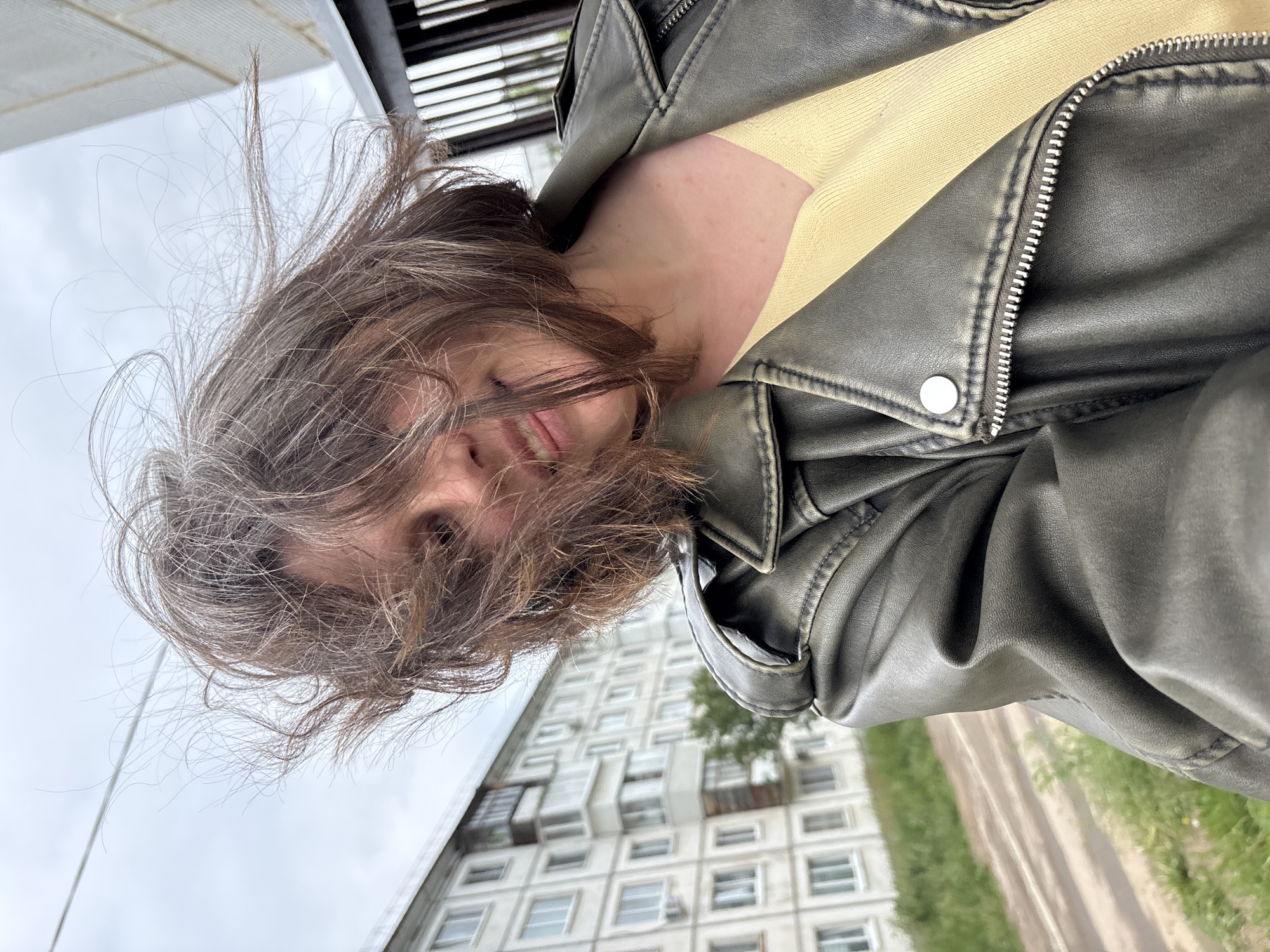 This media file is an outdoor portrait of a person, likely female, captured from a low angle with the image rotated 90 degrees clockwise. The person looks directly at the camera, their face partially obscured by their medium-brown, wind-blown hair, which also contains lighter, possibly grey strands. A slight smile is visible on their lips.
The individual is wearing a dark, distressed leather-look jacket, appearing to be dark grey or olive green, with a prominent silver zipper and a silver snap button detail on the lapel. A light yellow or beige ribbed top is visible underneath the jacket, around the neckline.
The background features a large, multi-story apartment building with numerous rectangular windows and some balconies, characteristic of Soviet-era or post-Soviet residential architecture. Below the building, there's a strip of green grass and a dirt or unpaved path. The sky is overcast and grey, indicating a cloudy daytime setting, with the wind visibly affecting the person's hair. No discernible text is visible in the image. The scene is set in Chaikovsky, Russia.